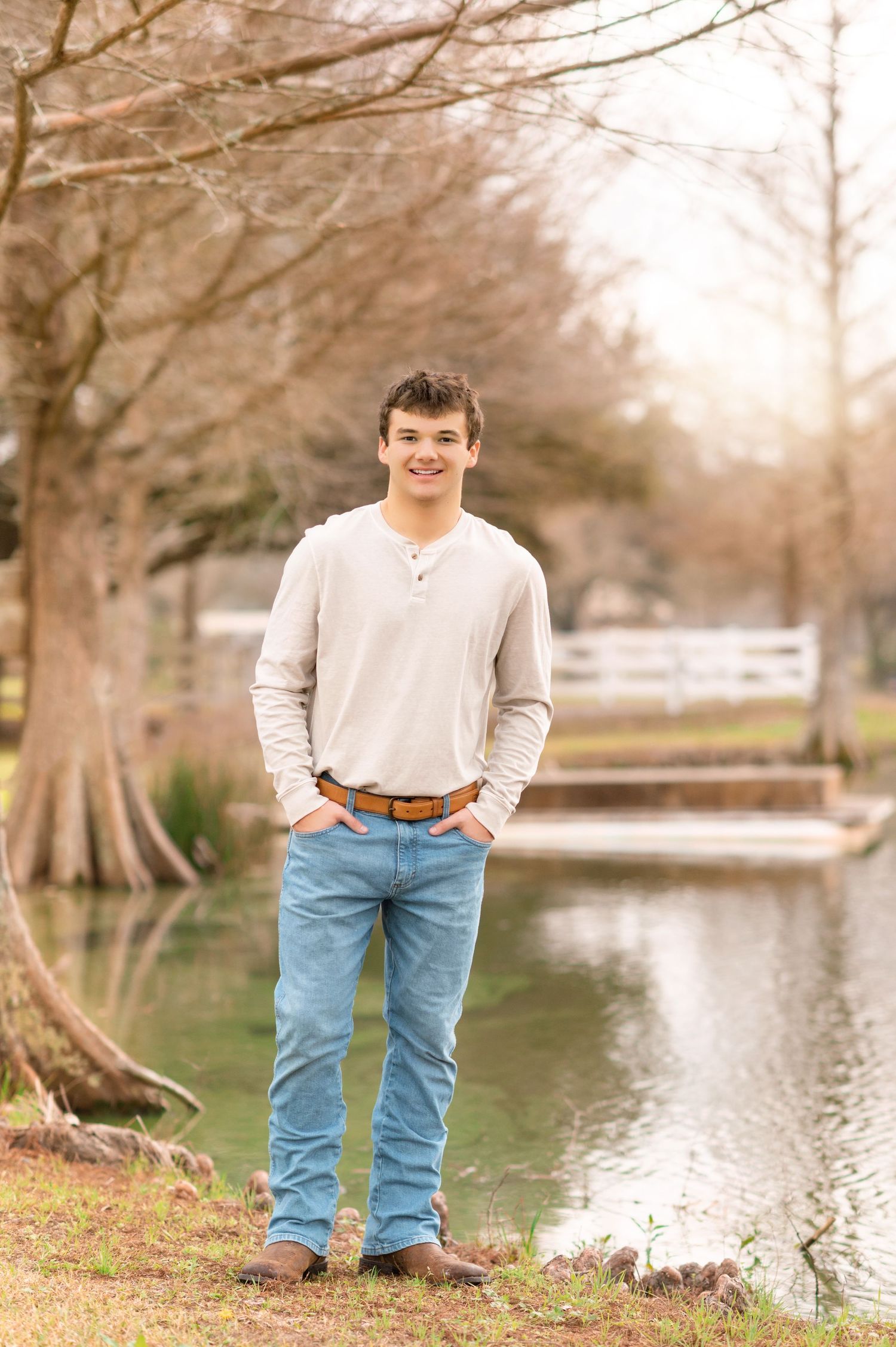 Young man in beige henley and jeans smiling outdoors near a pond with bare trees in a park setting.