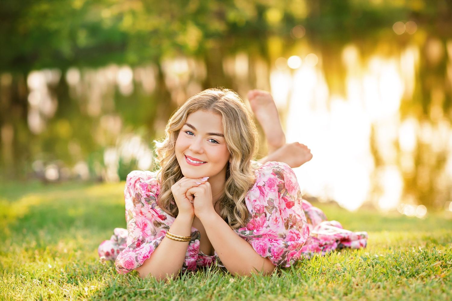 Smiling blonde teen girl in pink floral dress lying on green grass near a sunlit lake.