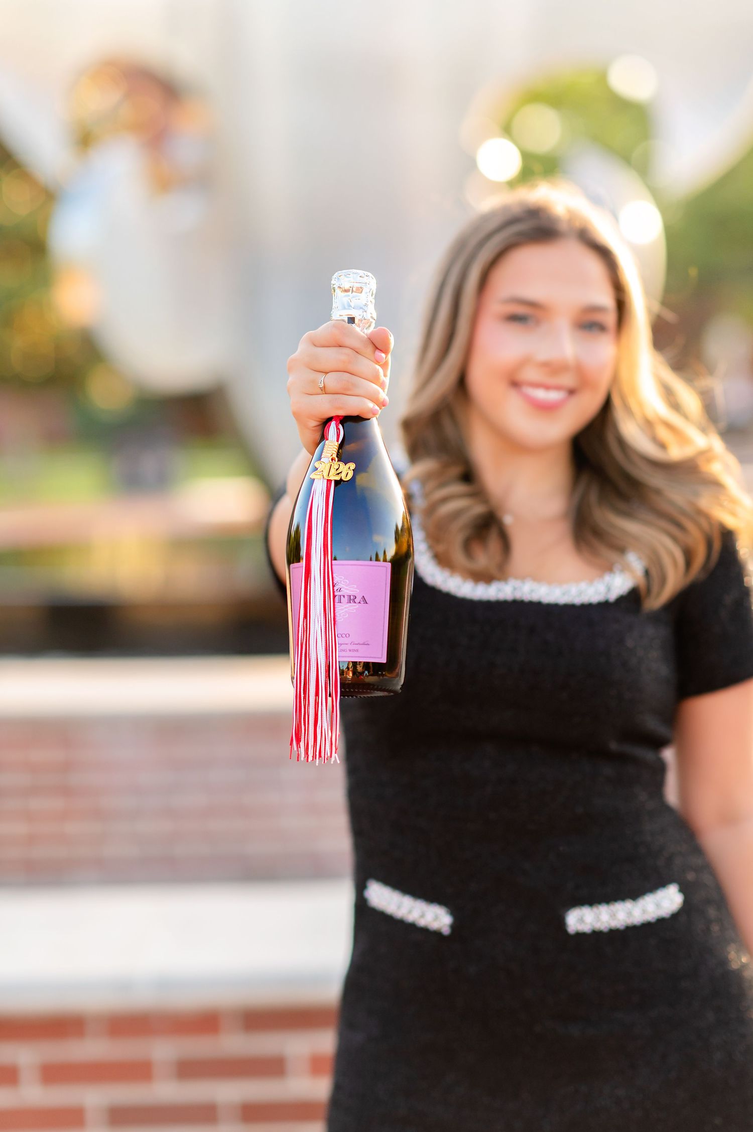 Smiling graduate holding champagne bottle with pink tassel decoration at outdoor celebration.