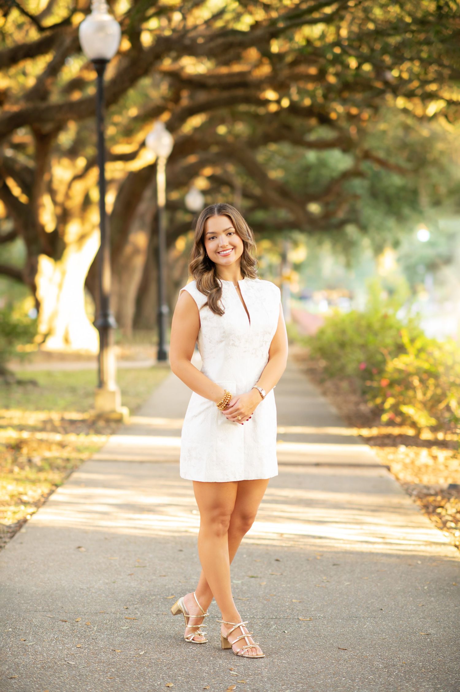 Young woman in white dress and nude heels posing on a tree-lined pathway in golden hour sunlight.