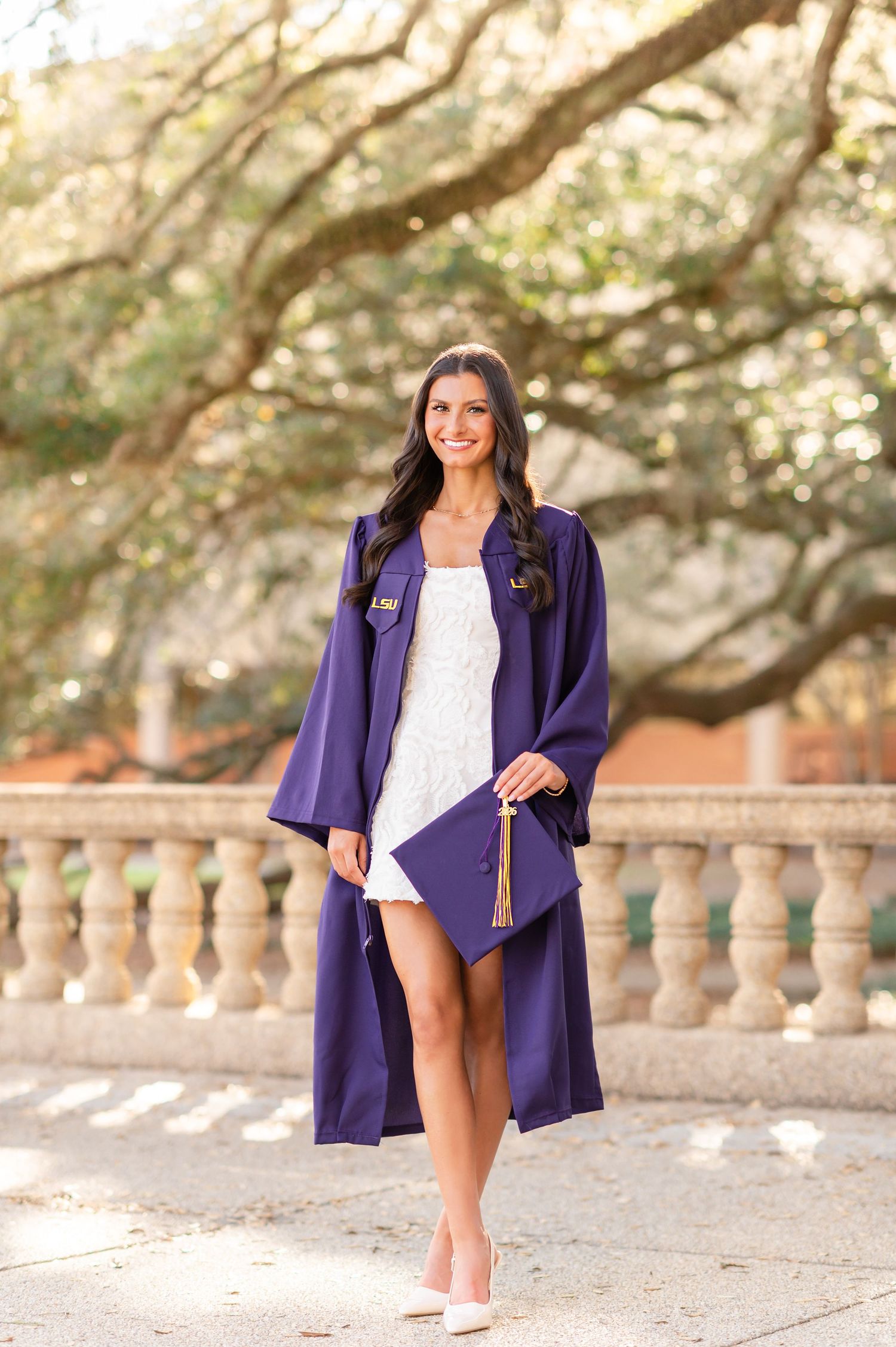 LSU graduate smiling in purple cap and gown over white dress, holding diploma cover outdoors.