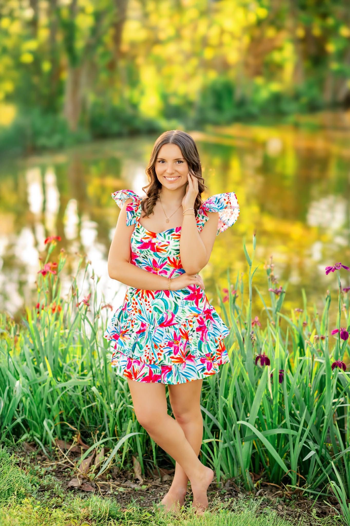 Young woman in colorful floral dress standing barefoot by a pond in a garden.