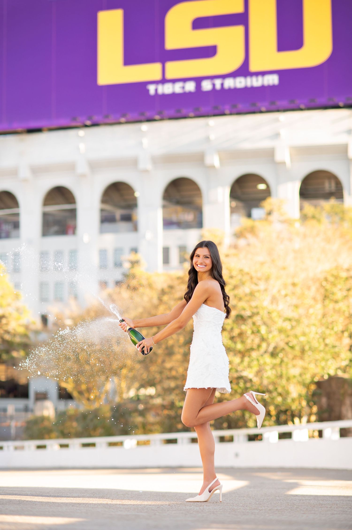Graduate in white dress pops champagne bottle in front of LSU Tiger Stadium on a sunny day.