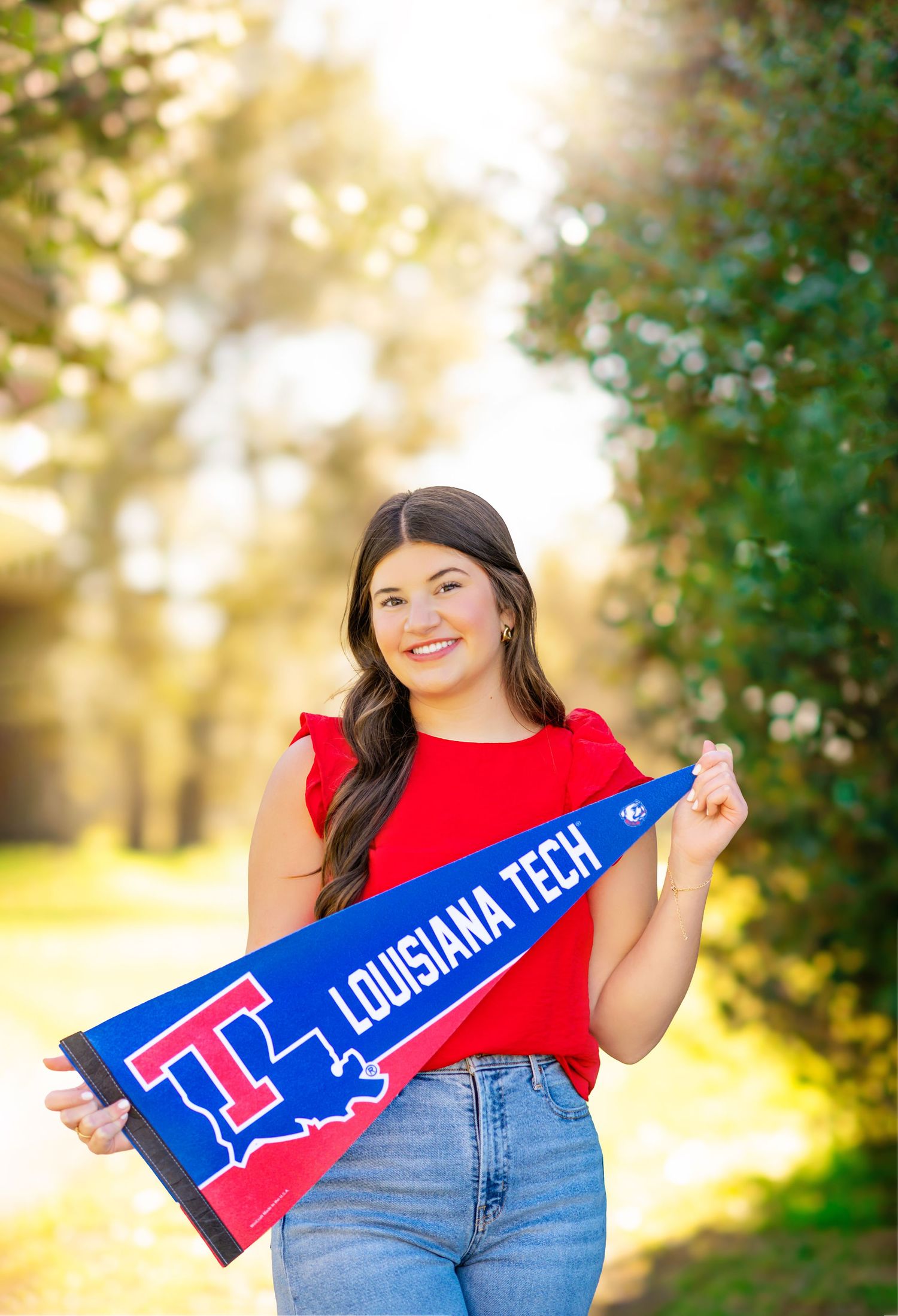 Smiling brunette woman in red top holding Louisiana Tech pennant flag outdoors in golden sunlight.