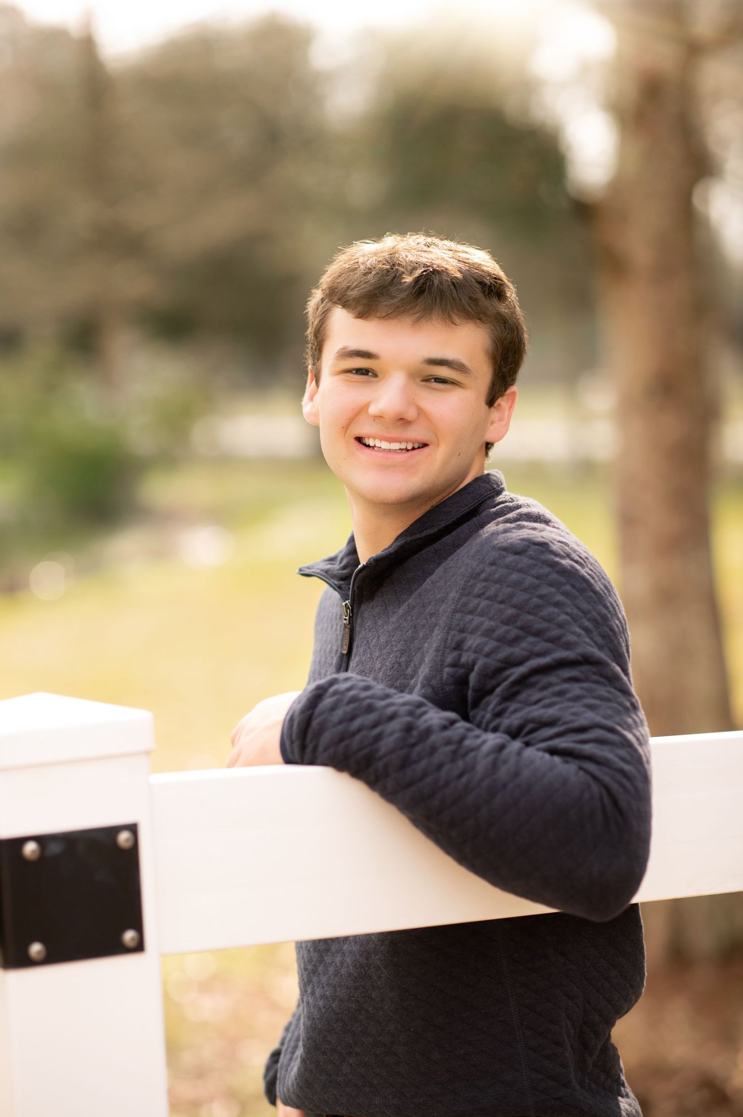 Smiling young man in navy quilted pullover leaning on white fence outdoors in a park setting.