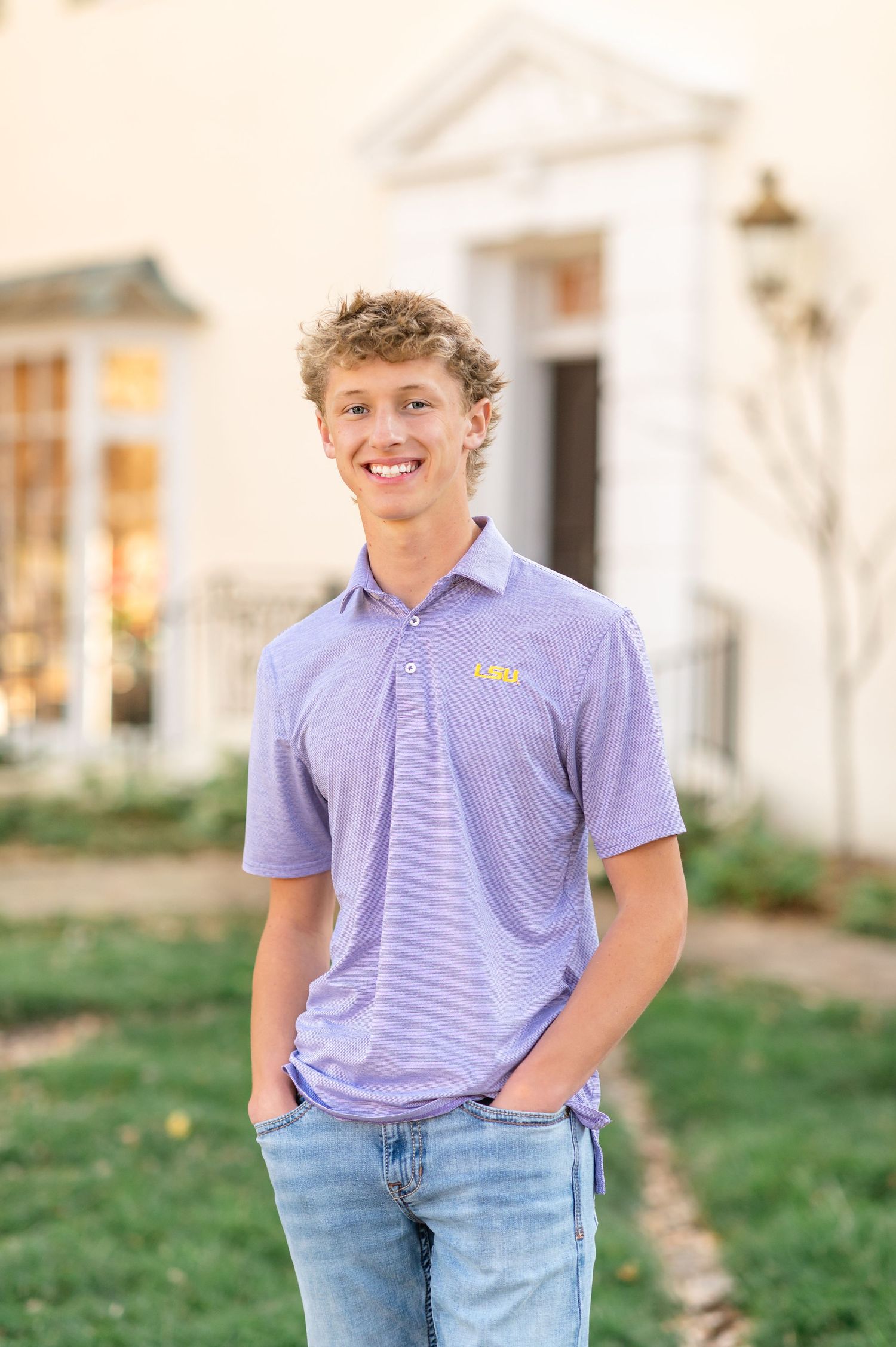 Smiling young man in LSU purple polo shirt and jeans posing outdoors in front of a white building.