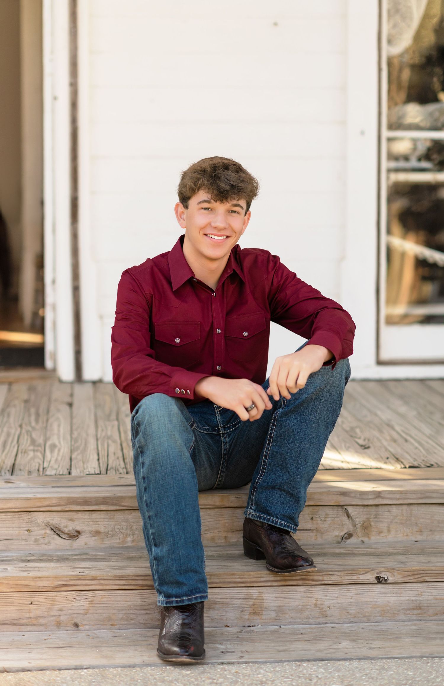 Smiling teen in maroon western shirt and jeans sitting on rustic wooden porch steps.