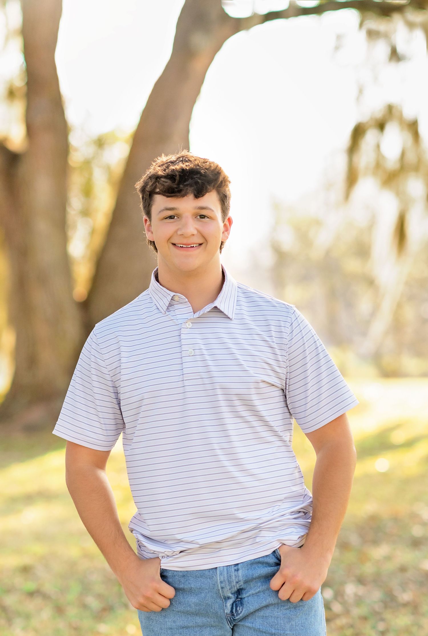 Smiling teenage boy in striped polo shirt posing outdoors near a large tree with Spanish moss.