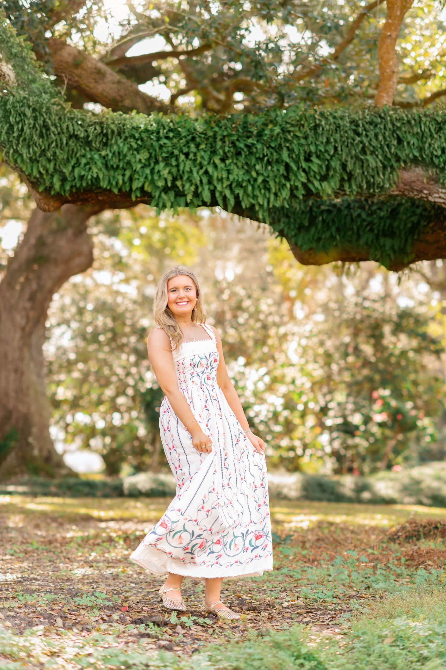 Young blonde woman in floral midi dress smiling under a large moss-covered oak tree in a lush garden.