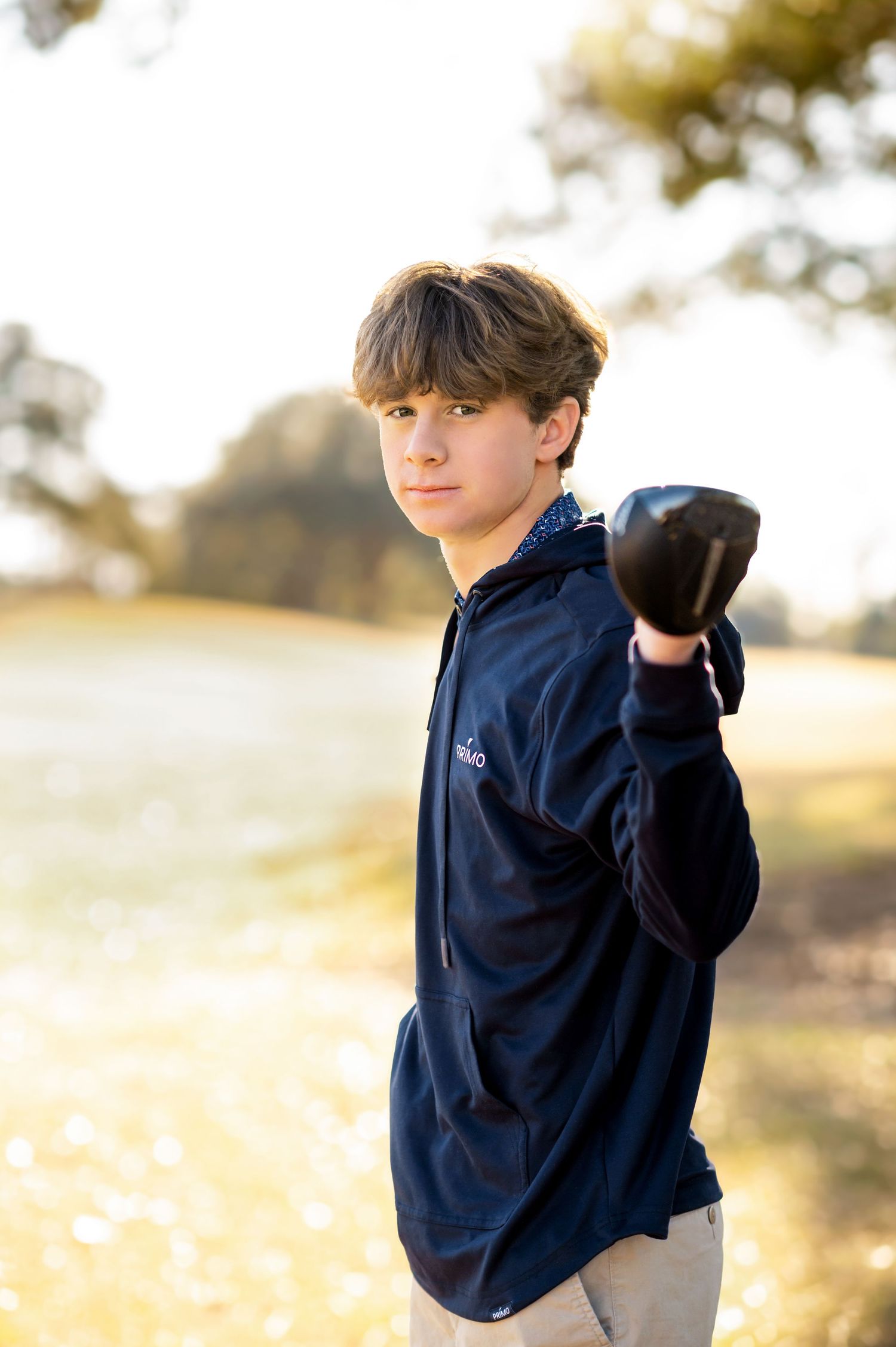 A teenage boy in a navy hoodie holds a golf club toward the camera outdoors in soft golden light.