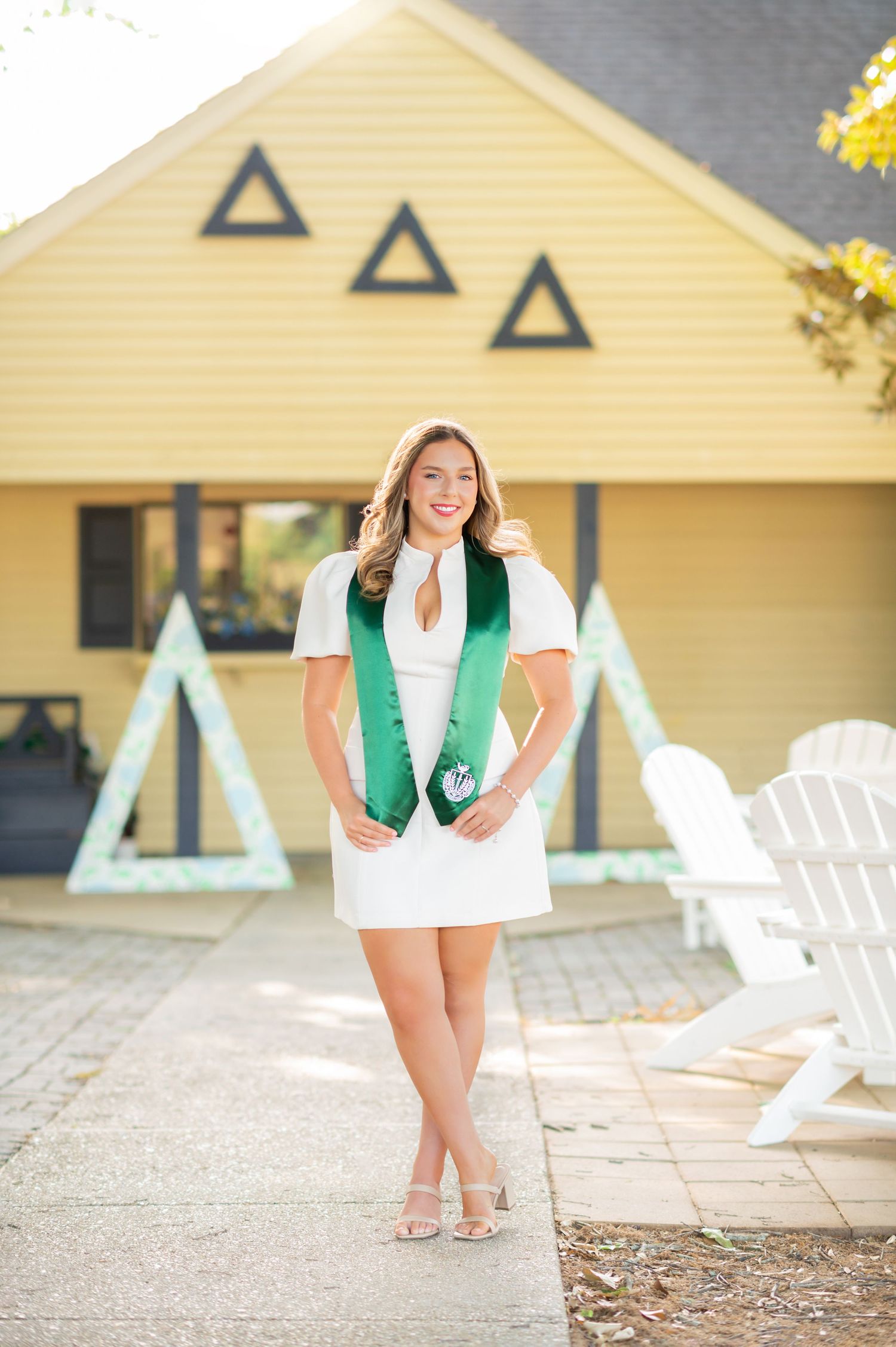 Young woman in white dress and green Delta Delta Delta graduation stole poses outdoors at sorority house.