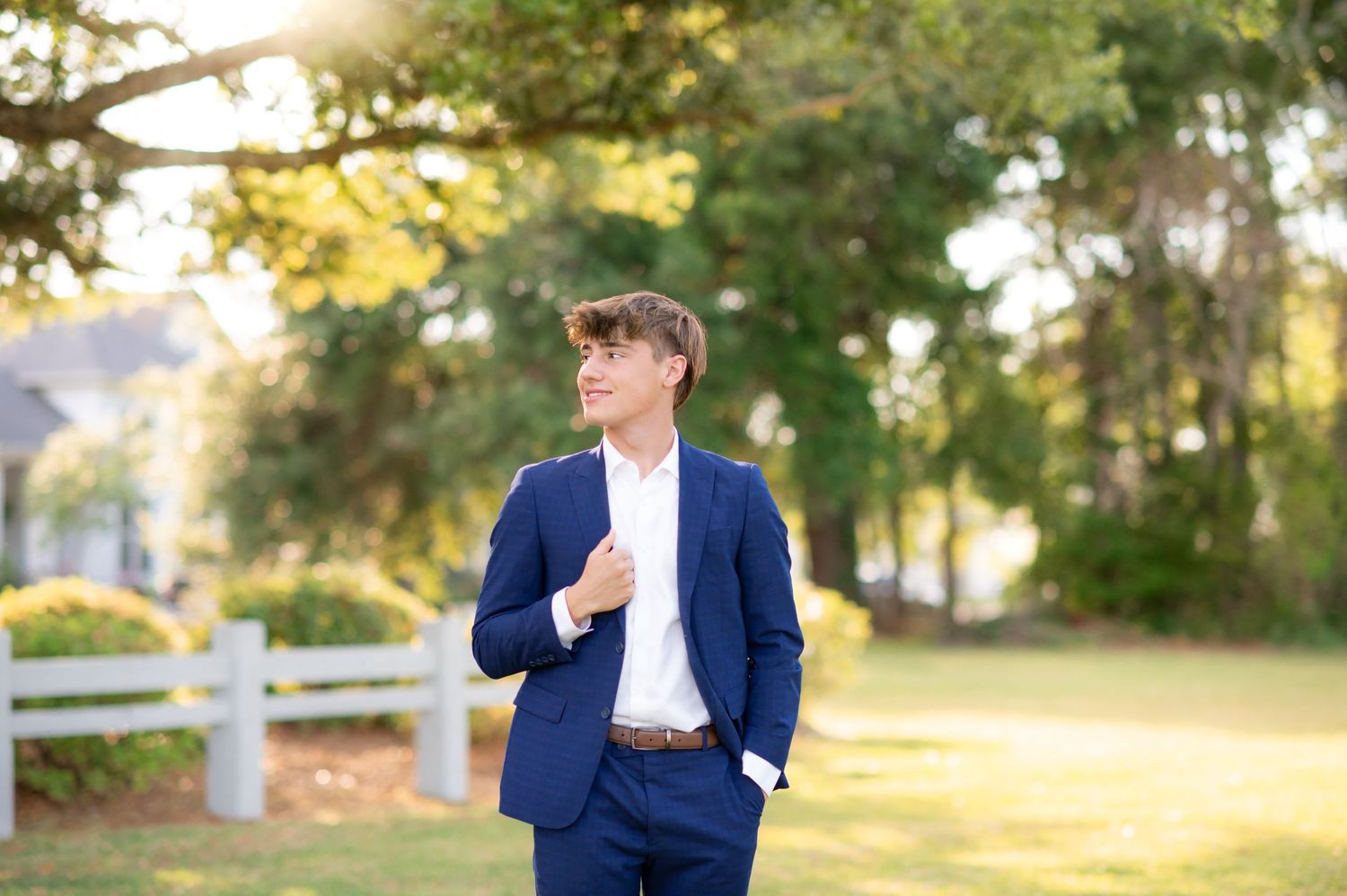 Young man in navy blue suit posing outdoors near white fence with lush green trees in sunlit background.