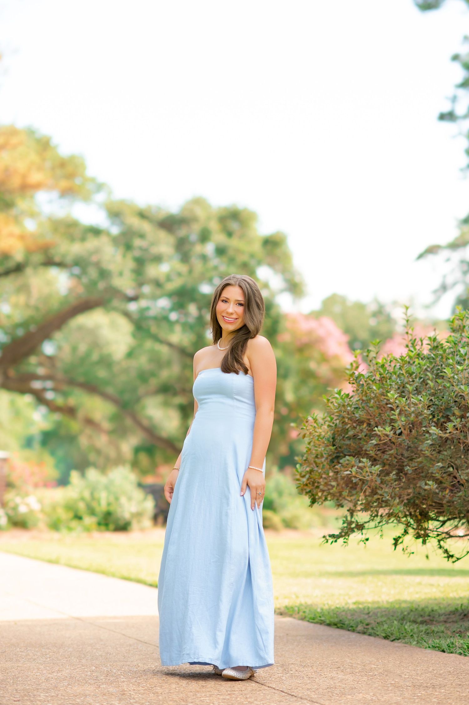 Pregnant woman in a light blue strapless maxi dress poses on a garden path surrounded by lush greenery.