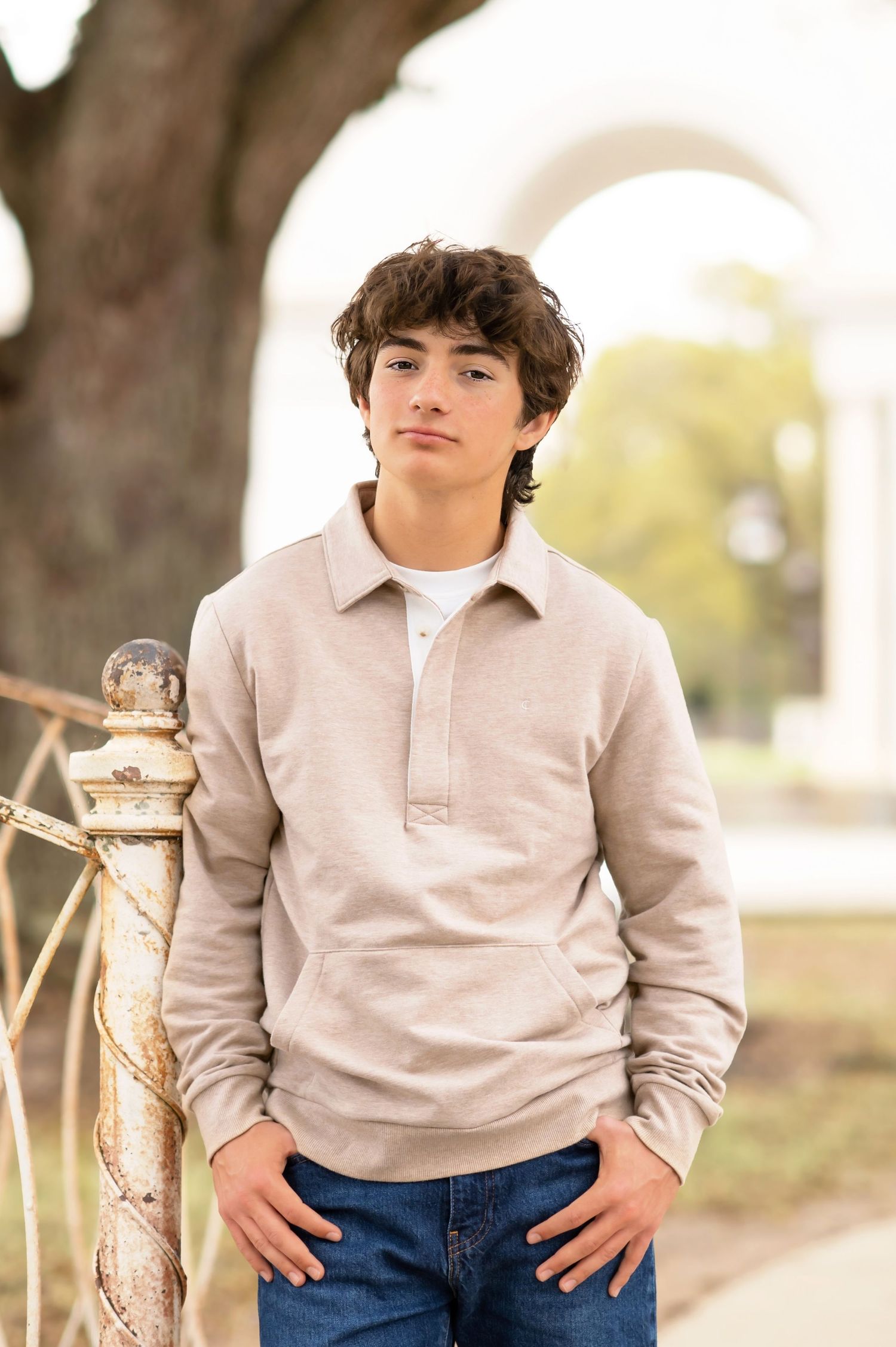 A teenage boy in a beige pullover and jeans poses confidently near a decorative fence outdoors.