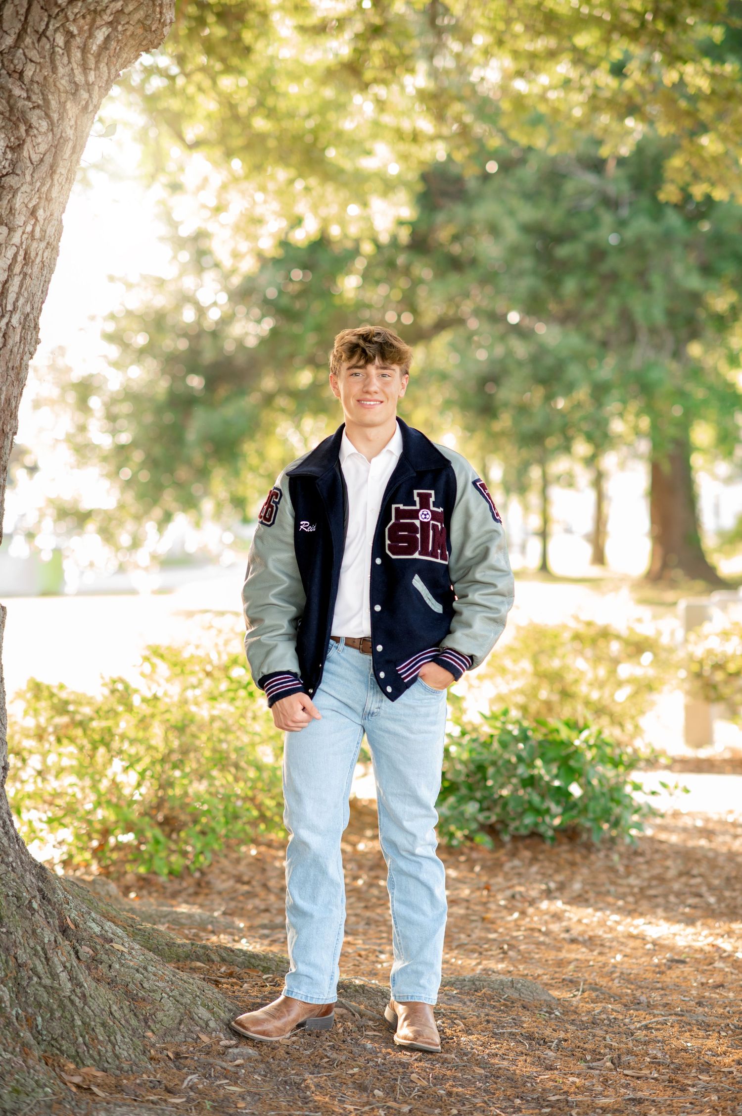 Young man in letterman jacket and jeans posing outdoors under trees for senior portrait photo.