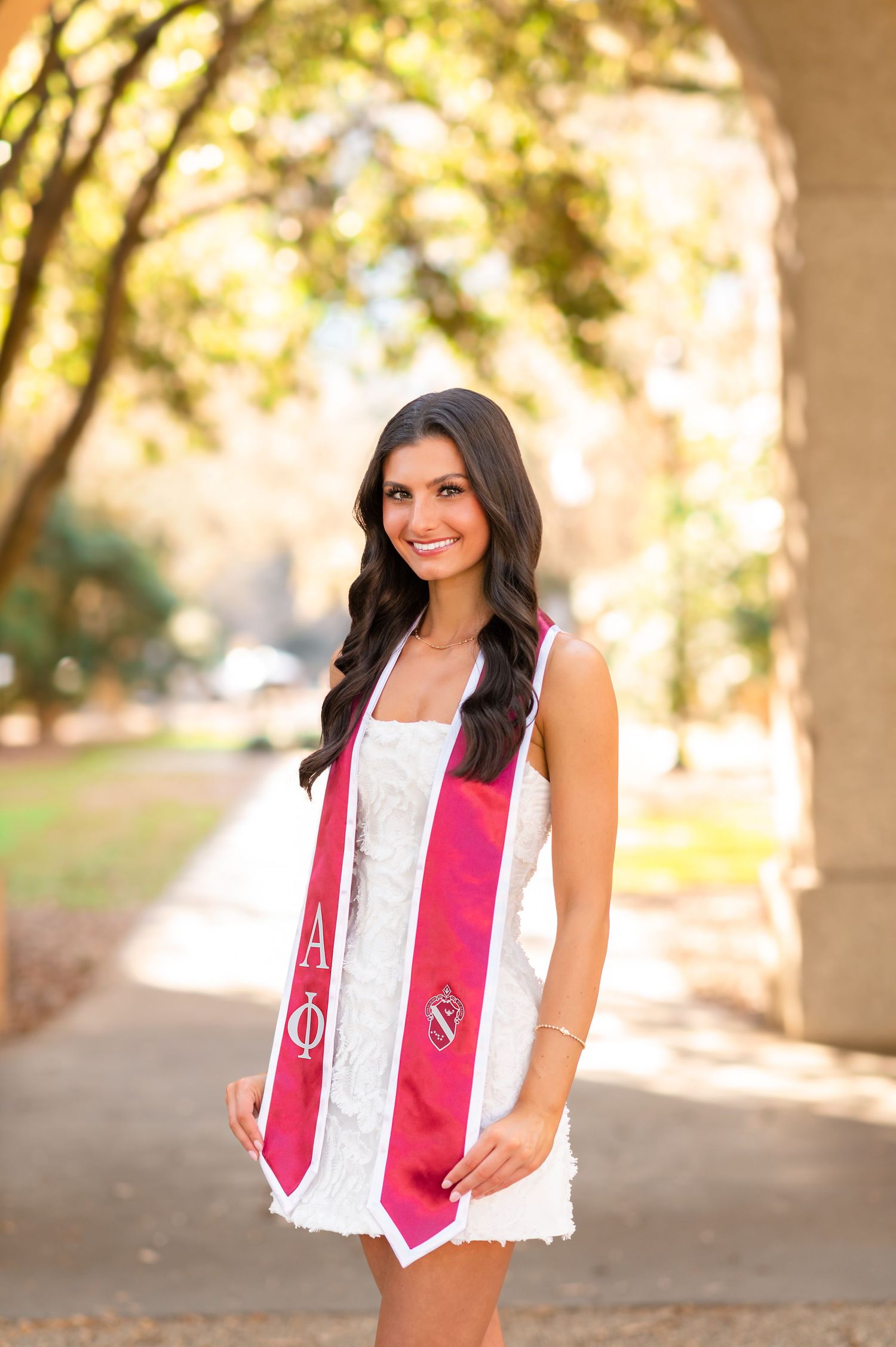 Young woman in white dress and pink Alpha Phi graduation stole smiling outdoors under archway.