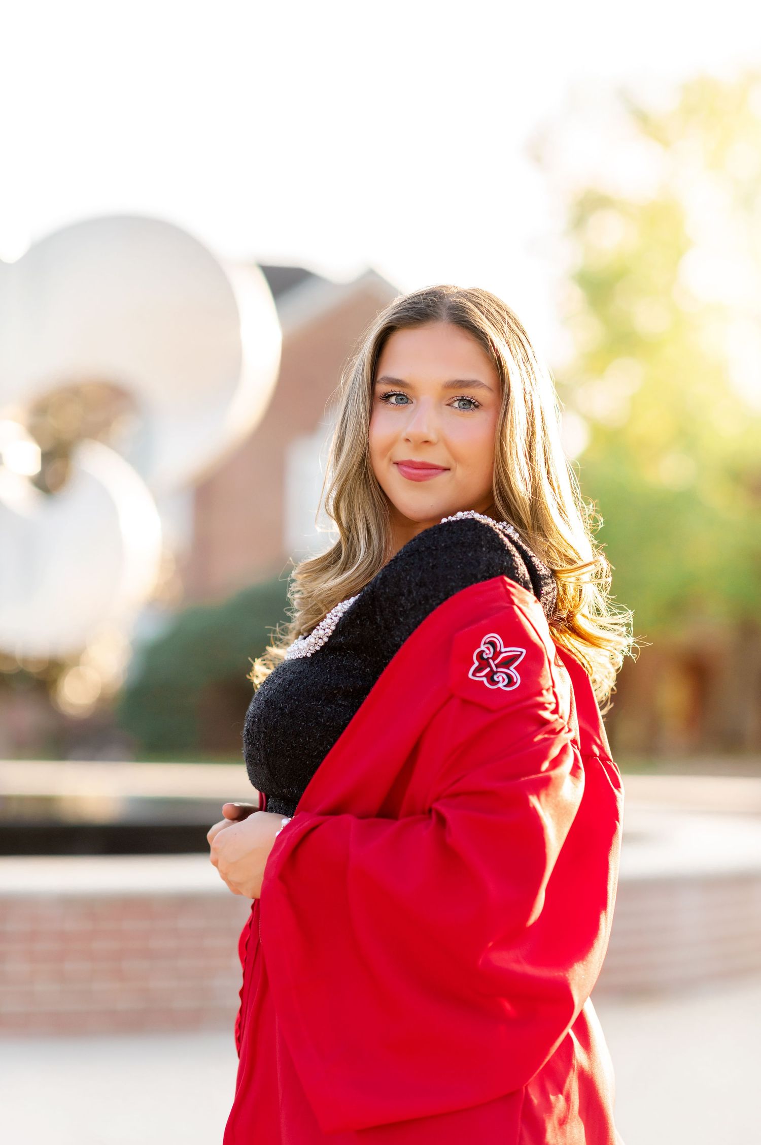 Smiling young woman in red graduation gown poses near large fleur-de-lis sculpture on sunny campus.
