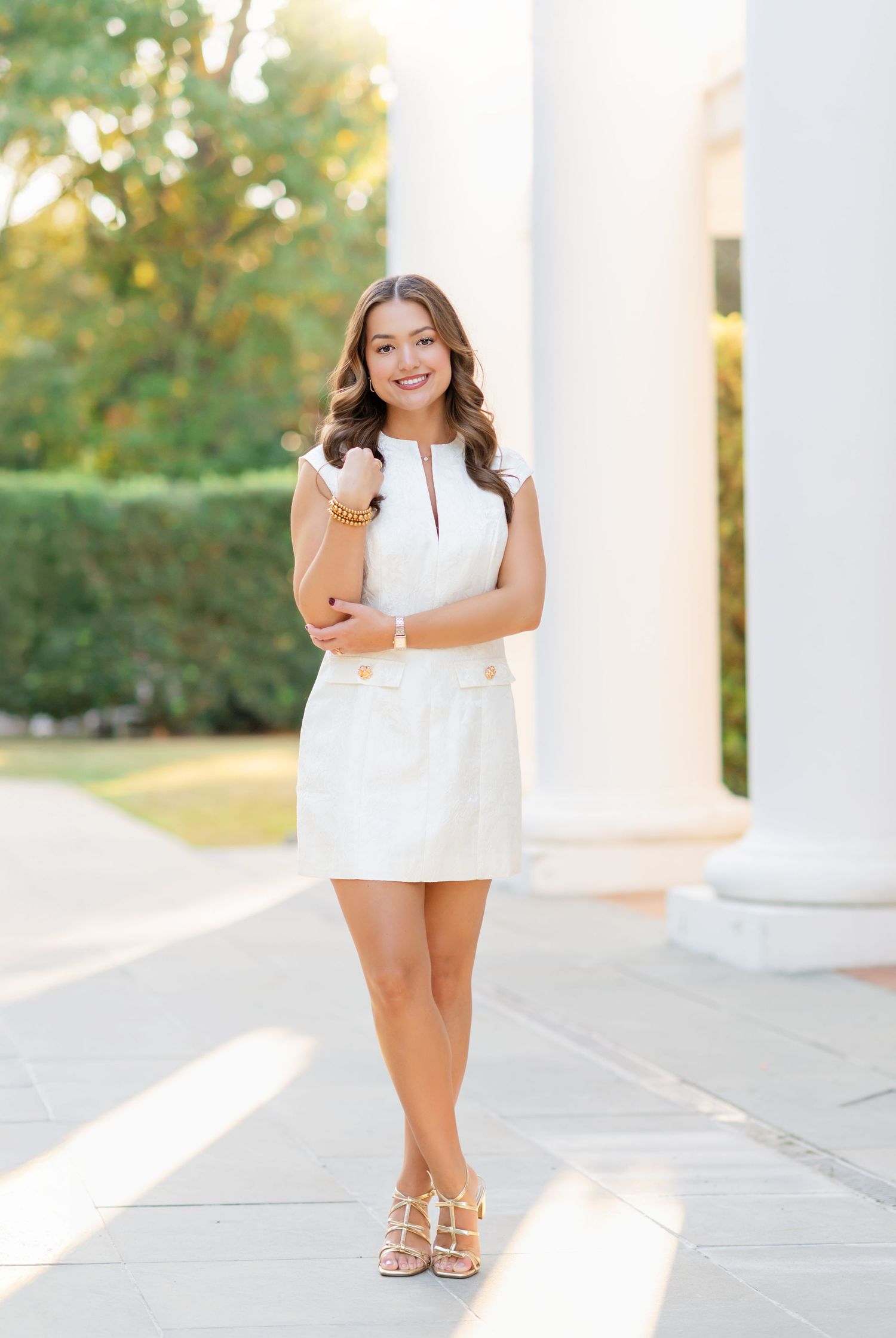Young woman in white mini dress posing outdoors near white columns on sunny day.