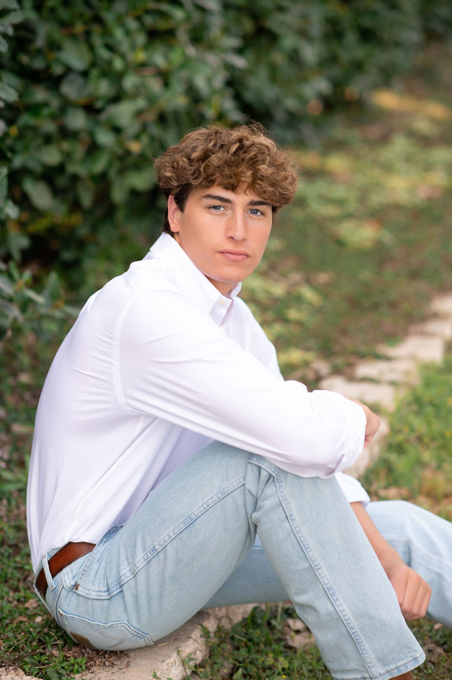 Young man with curly hair in white shirt and jeans sitting outdoors against lush green ivy backdrop.