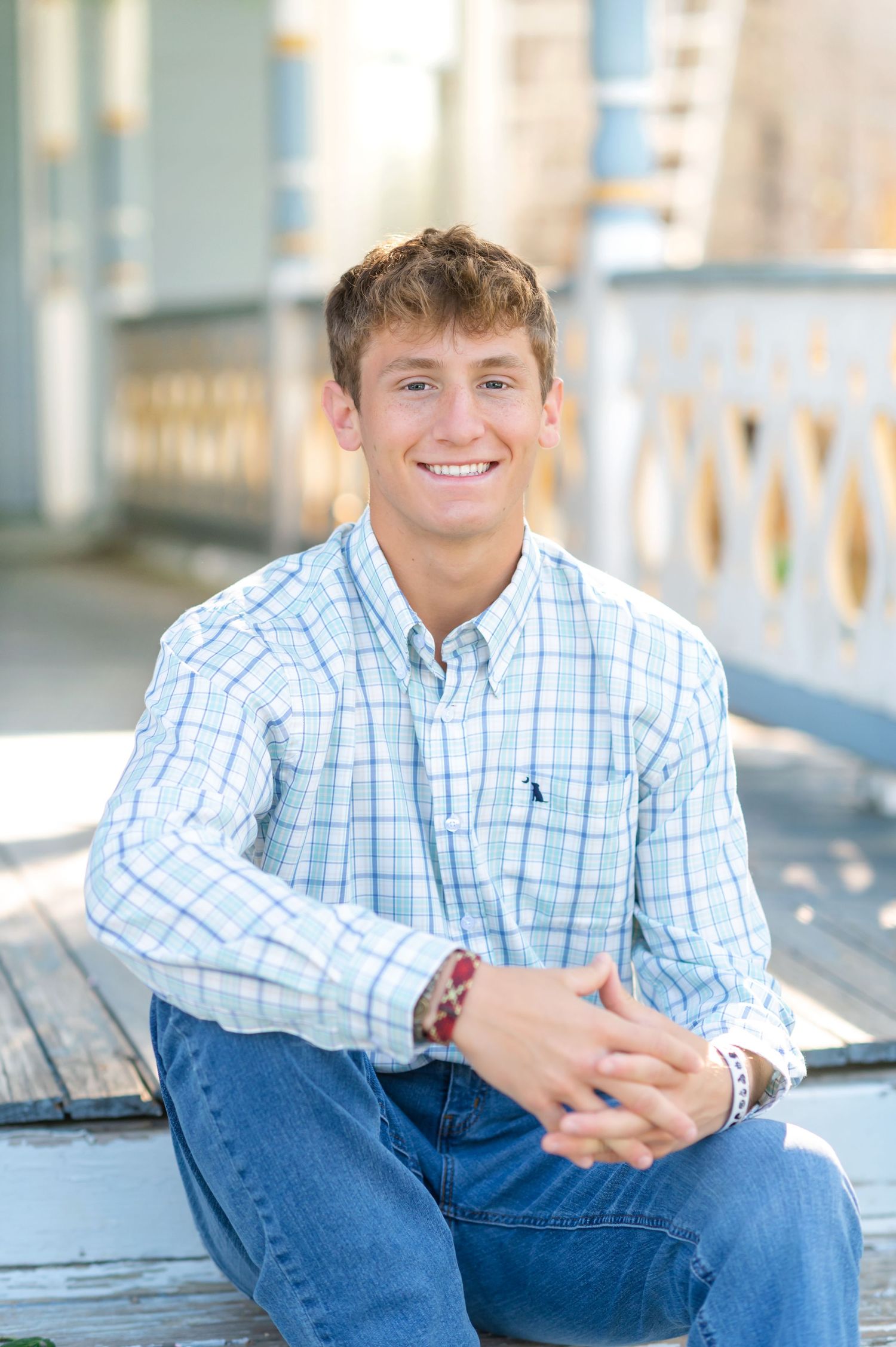 Smiling young man in plaid shirt and jeans sitting outdoors on wooden steps with hands clasped.