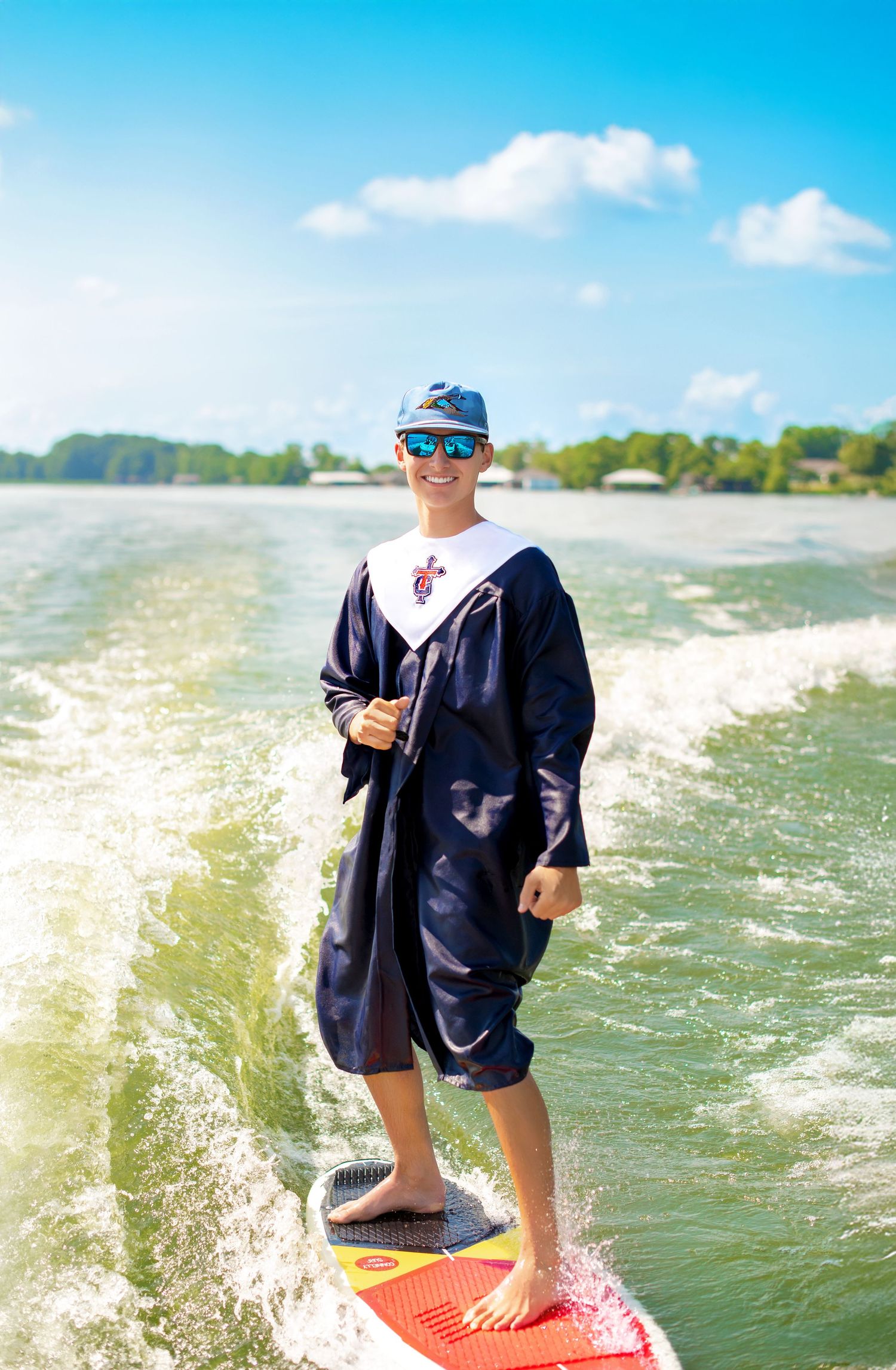 Young man wake surfing in graduation gown and cap smiling on sunny day on lake.