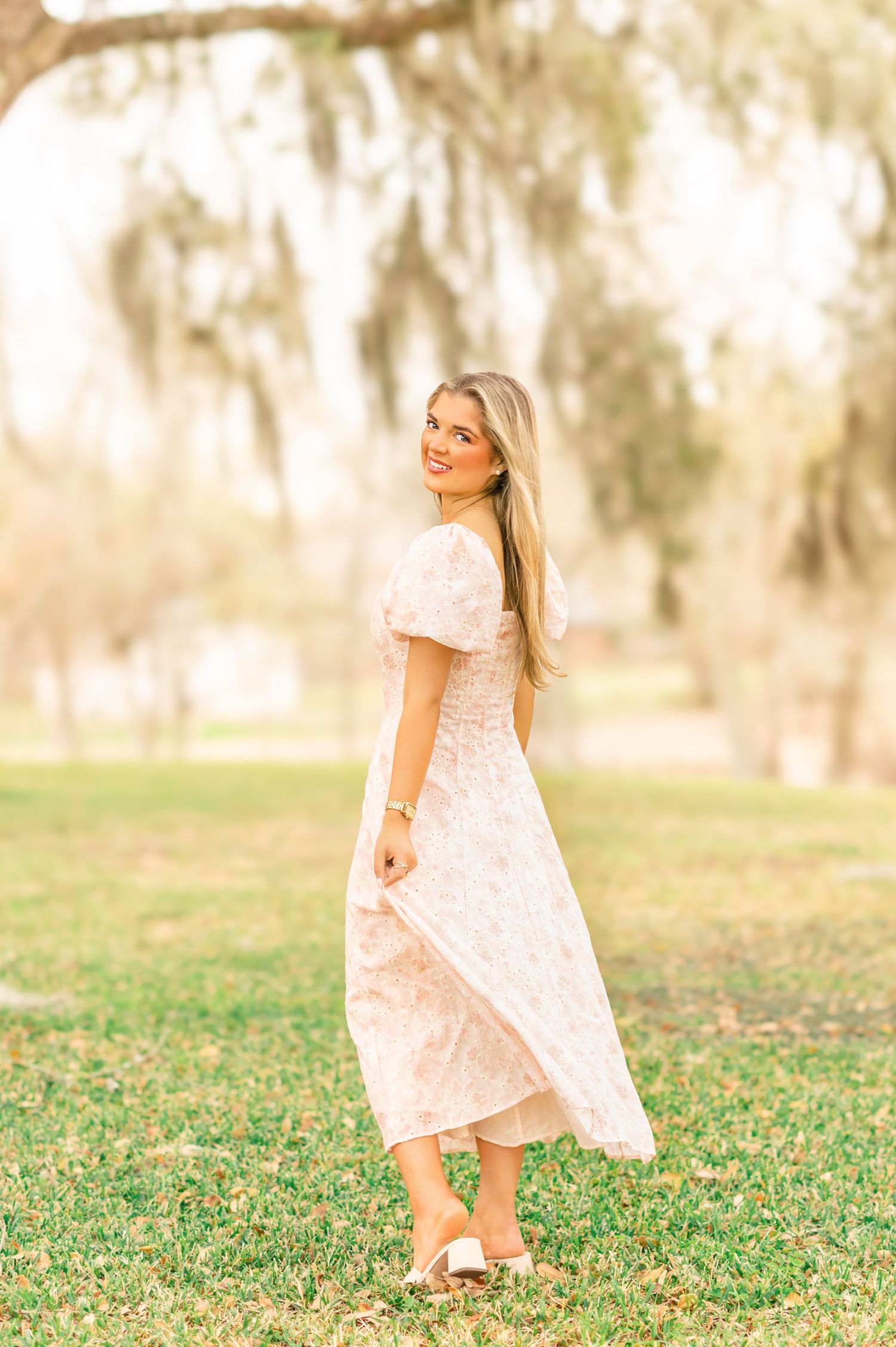 Smiling blonde woman in light pink floral midi dress twirling in sunny outdoor park with Spanish moss trees.