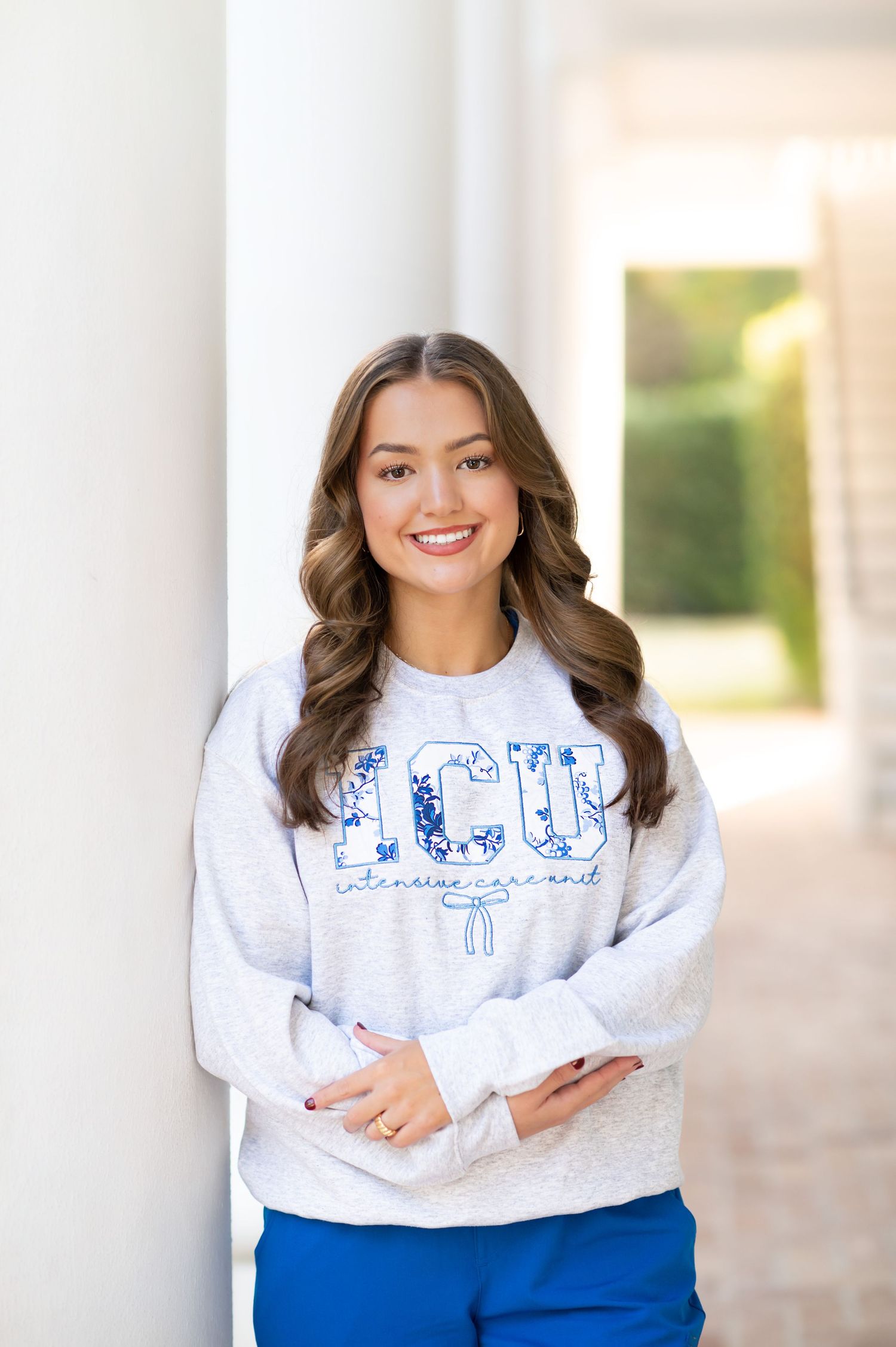 Smiling young woman in ICU sweatshirt and blue pants leaning against white column outdoors.