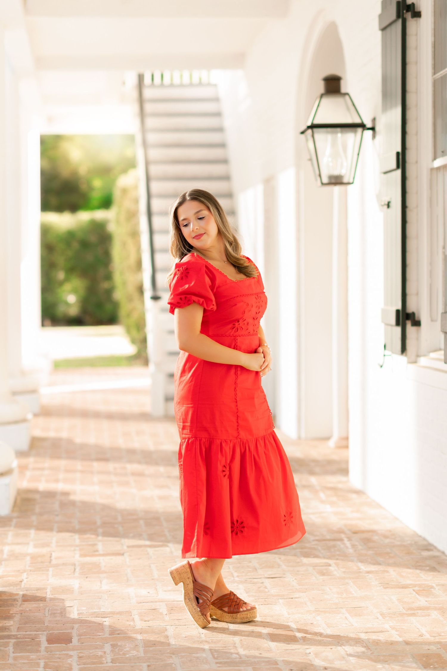 Woman in red midi dress with puff sleeves and wooden clogs posing in a sunlit white colonial corridor.