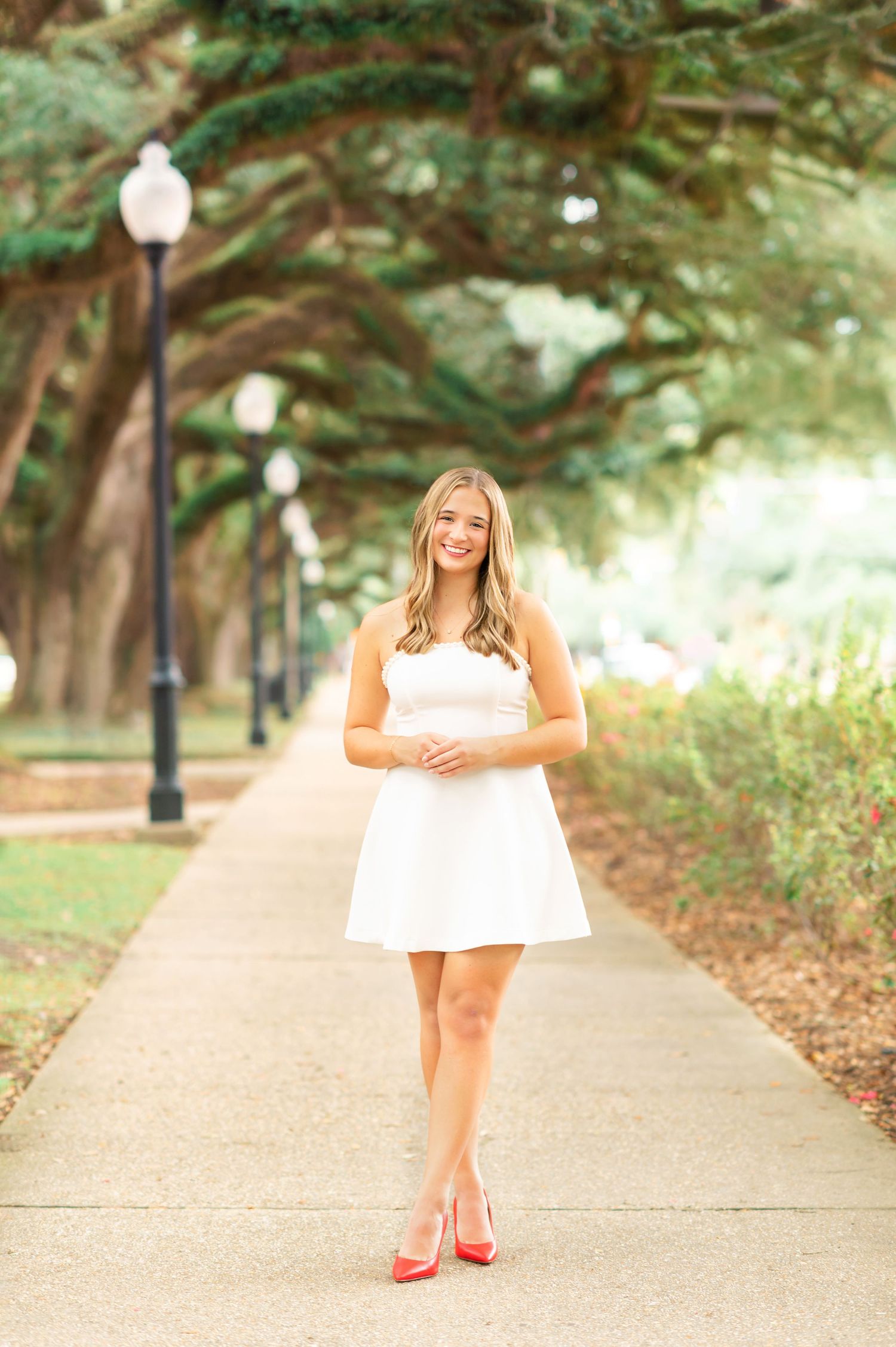 Young woman in white dress and red heels smiling on a tree-lined pathway in a sunny park.