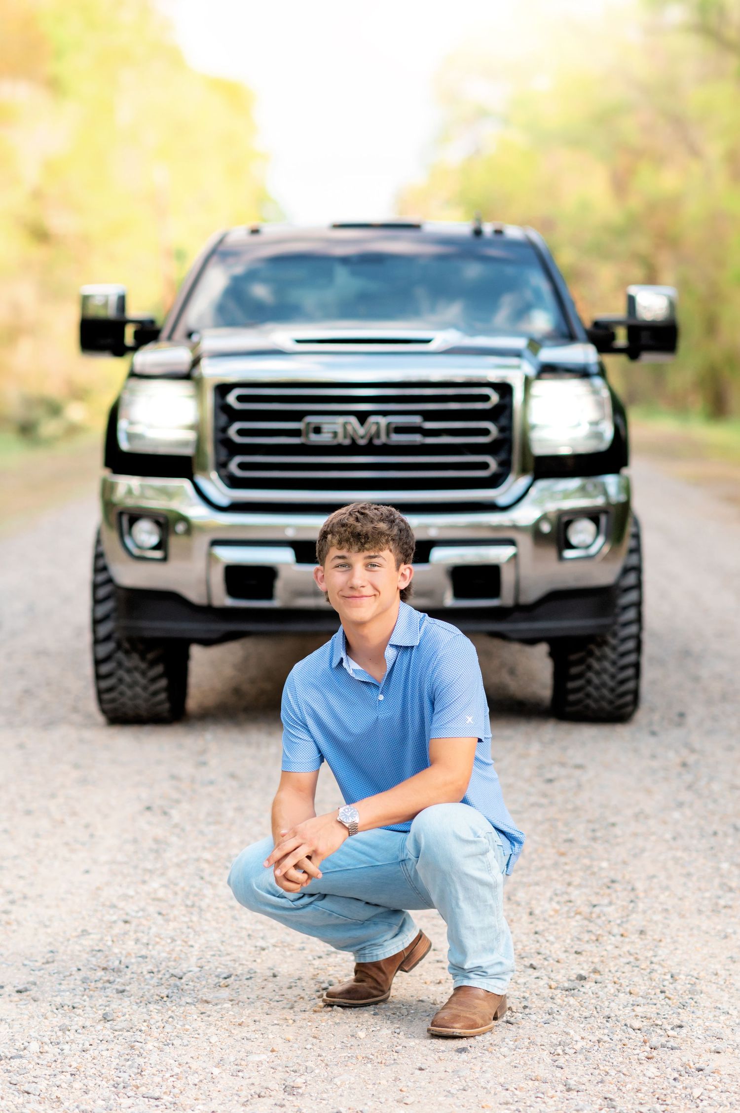 Teen boy in blue polo crouching in front of a lifted GMC truck on a gravel road for senior photos.