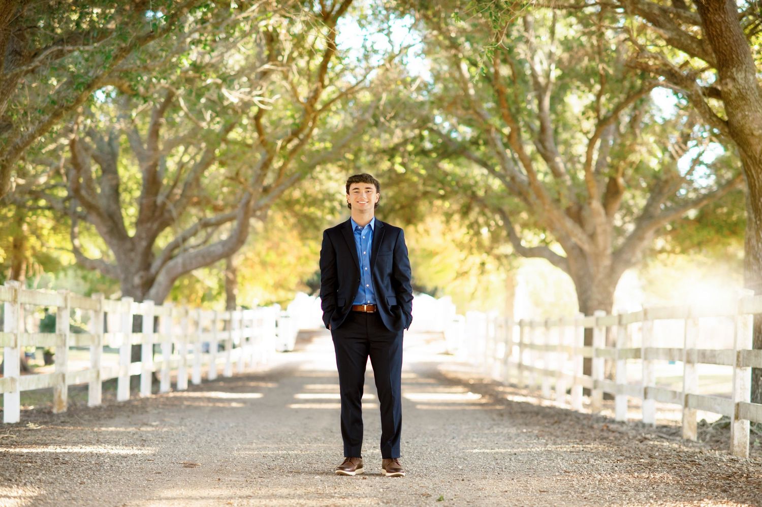 Young man in navy suit standing on a tree-lined gravel path with white fences, outdoor portrait photography.