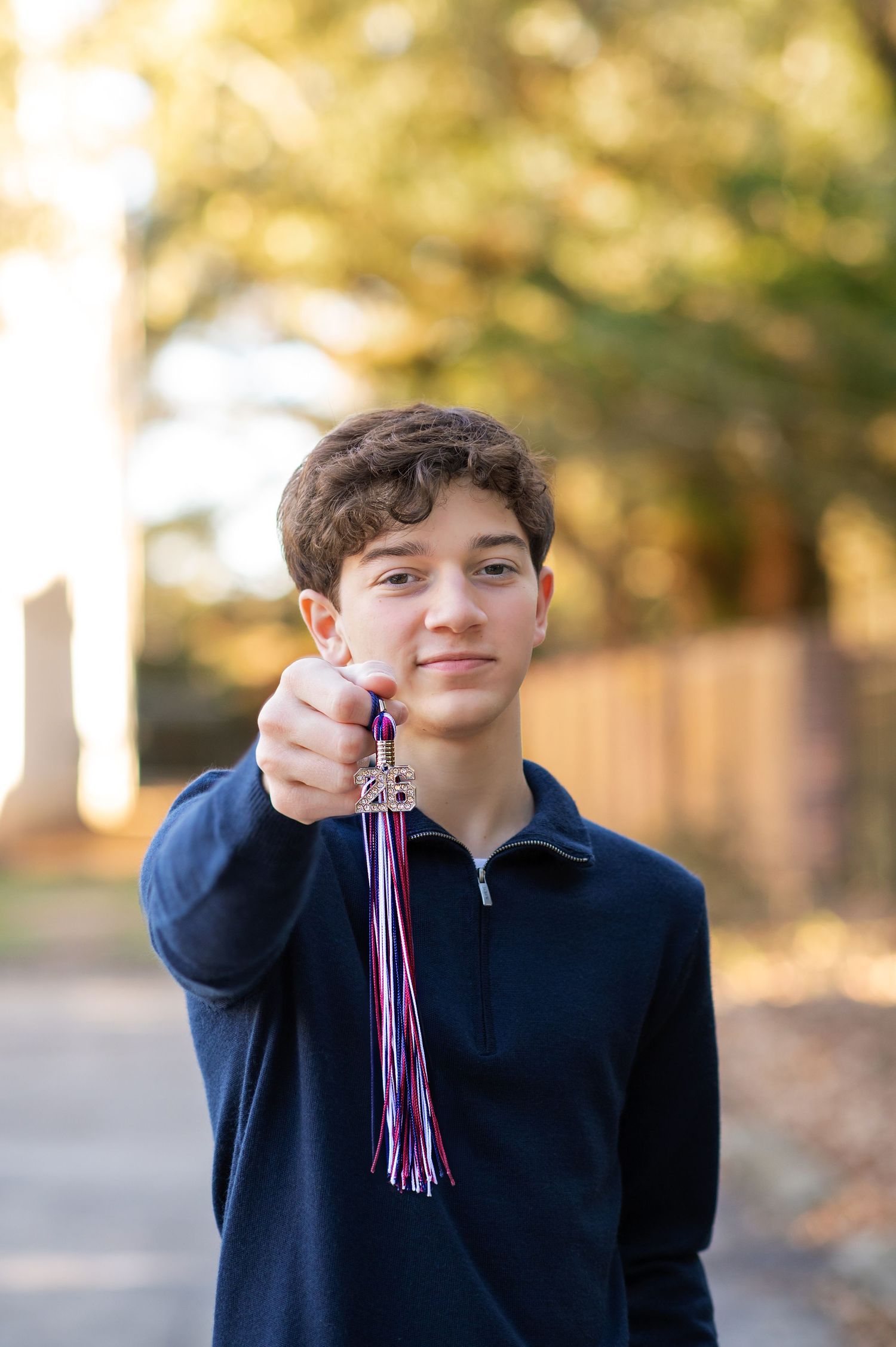 Teen boy in navy blue jacket holding a decorative charm with purple and pink ribbons outdoors.