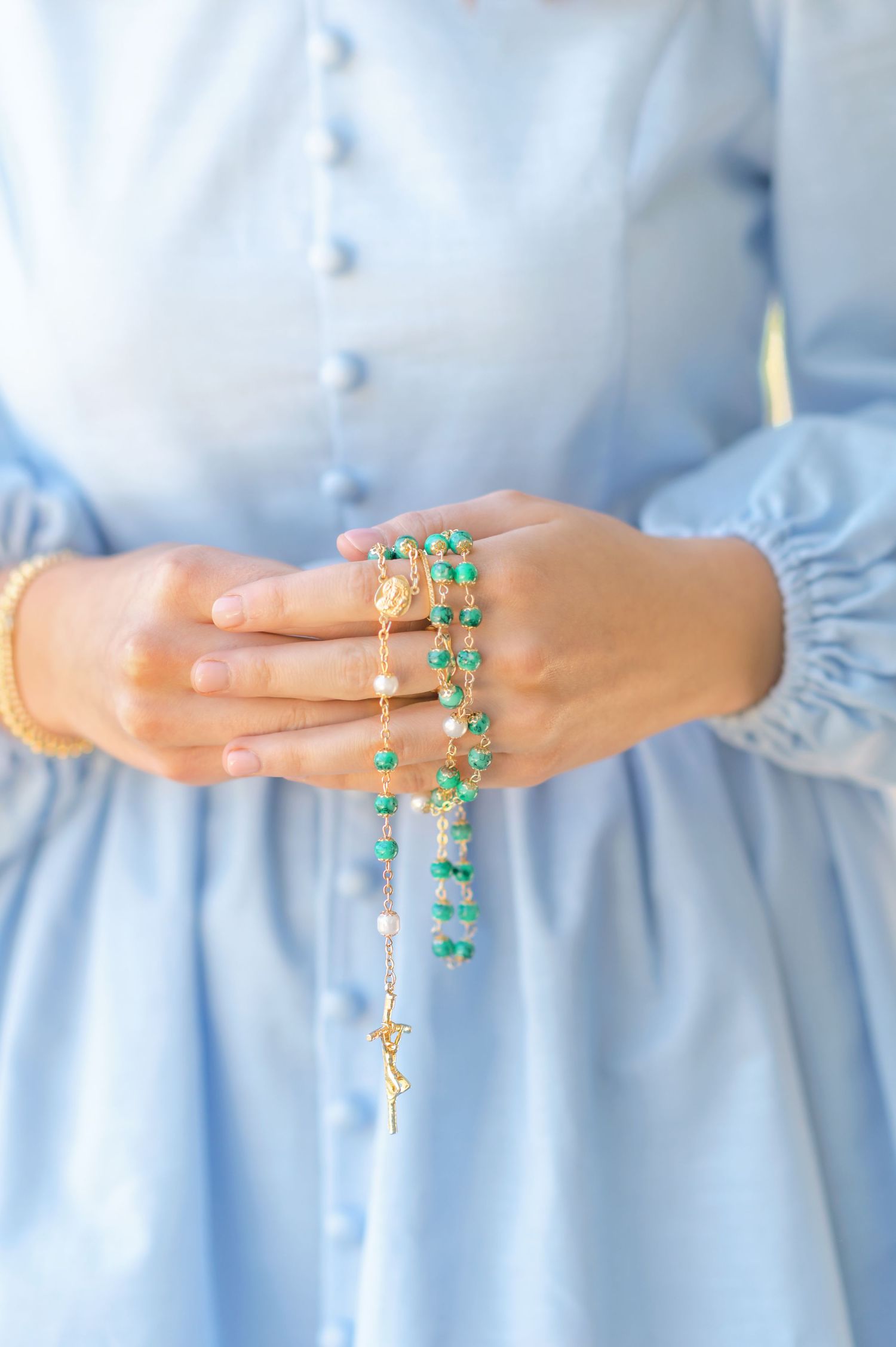 Close-up of hands holding a green and gold rosary against a light blue button-down dress.