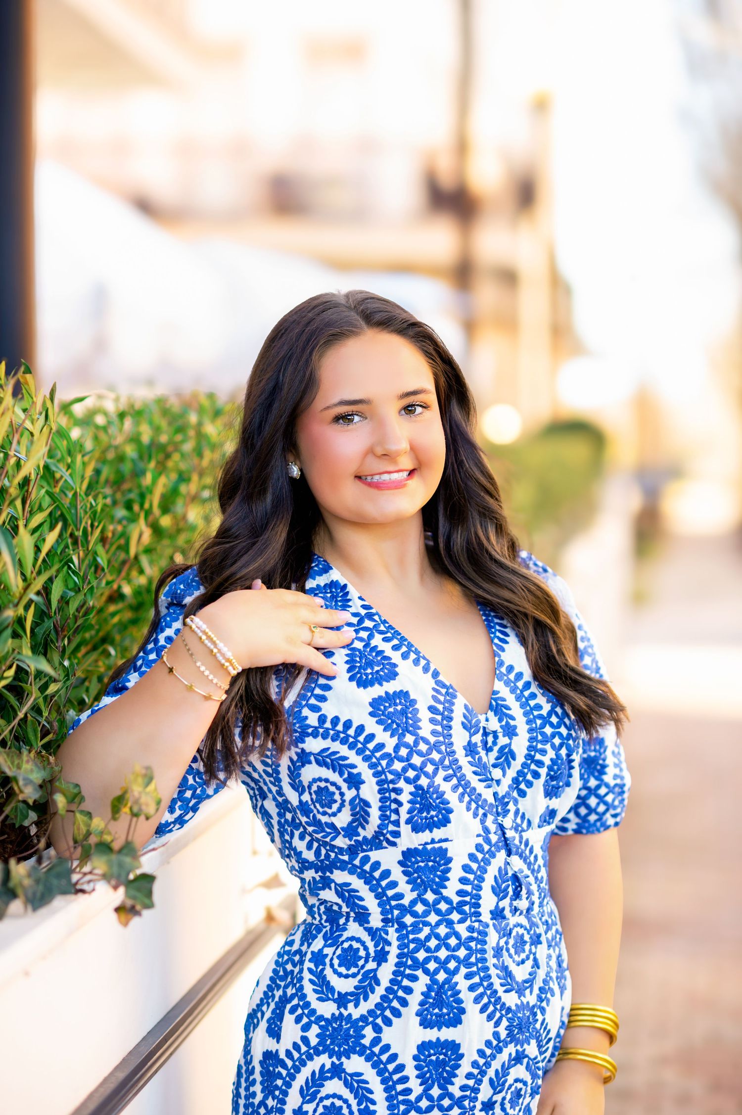 Brunette teen girl in blue and white patterned dress smiles outdoors near green hedges at golden hour.