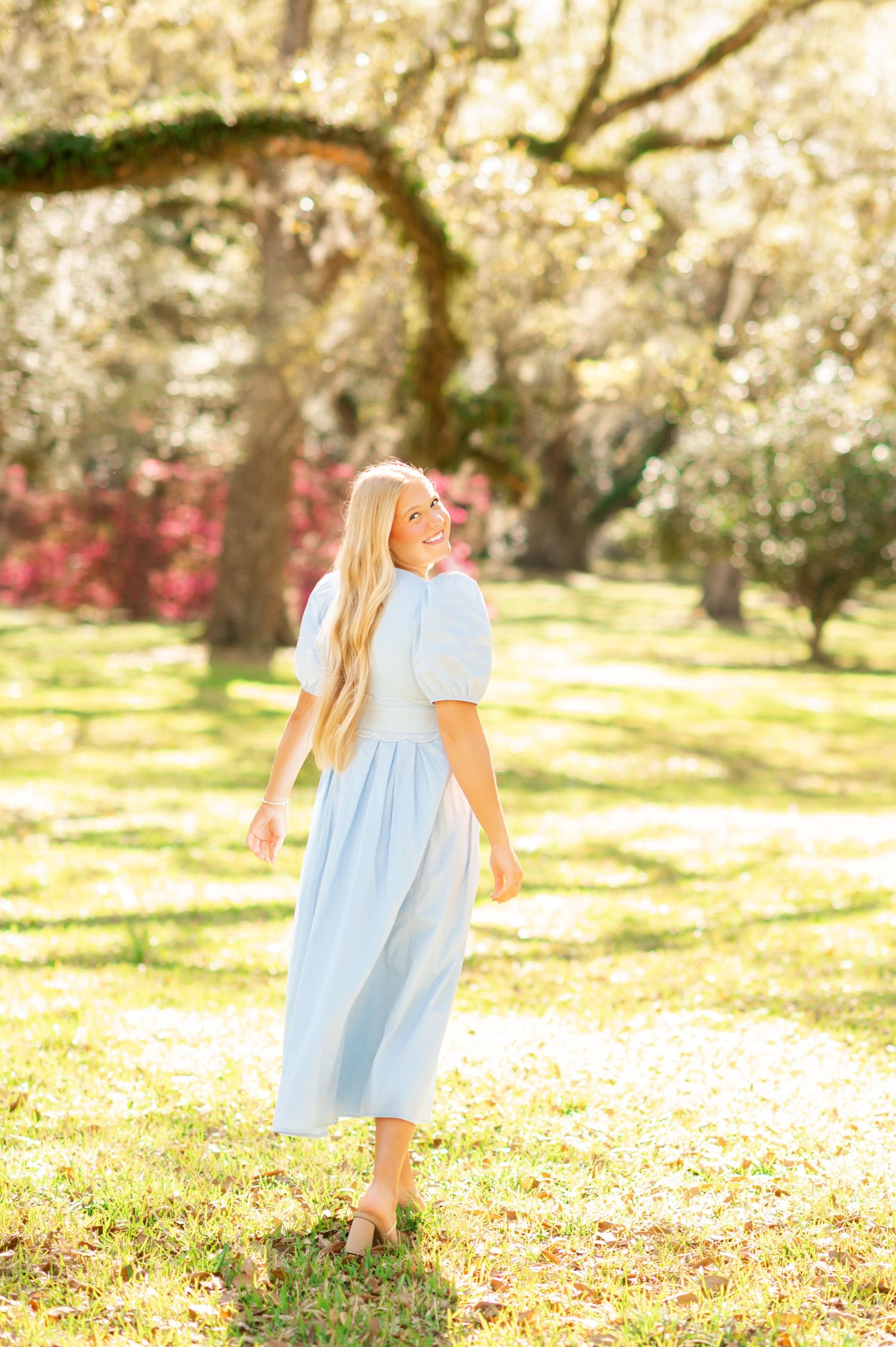 Blonde woman in light blue midi dress walks through sunlit park with moss-covered oak trees.