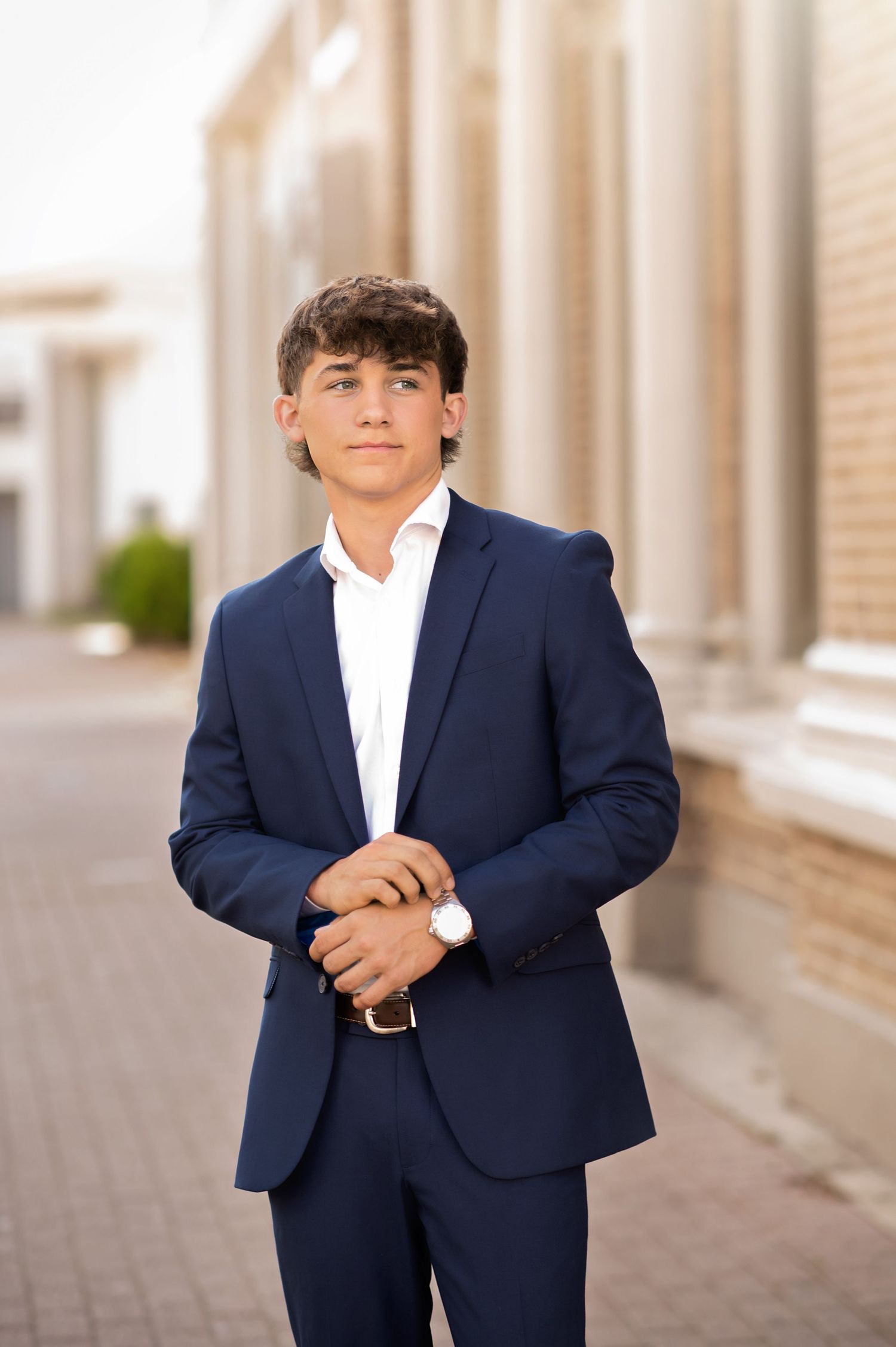 Young man in navy blue suit and white shirt posing outdoors near classical architecture columns.