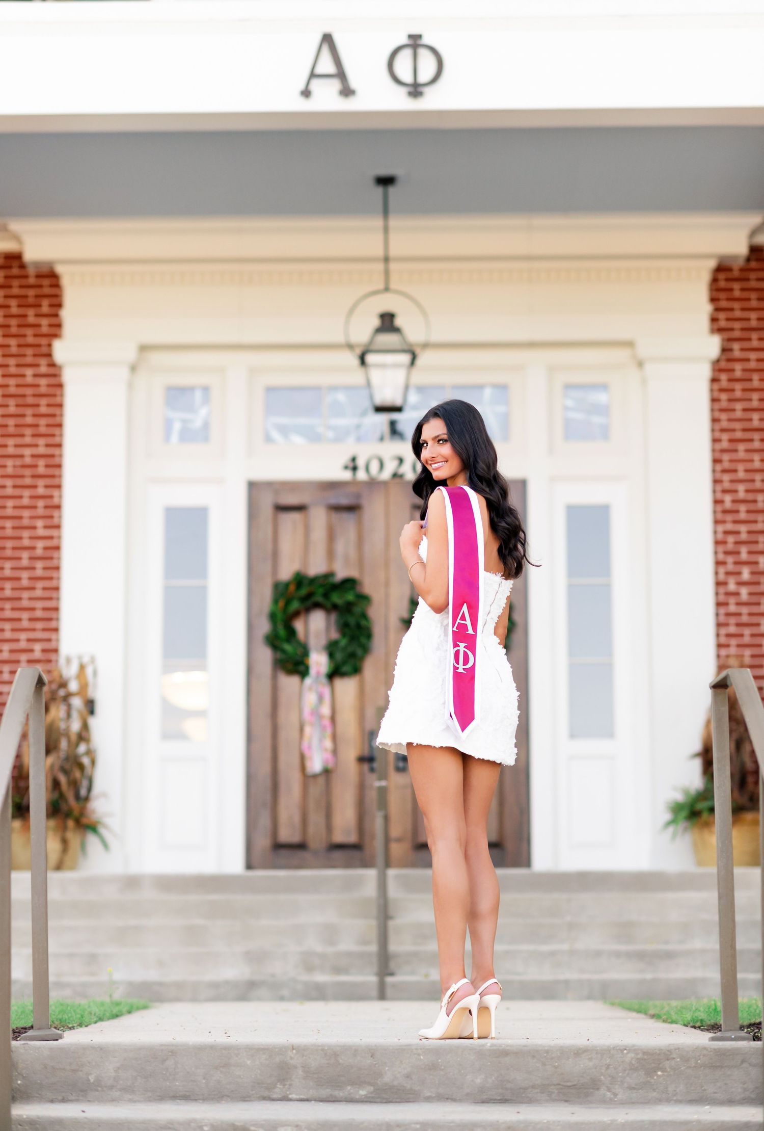 College woman in white dress with Alpha Phi graduation sash poses on sorority house steps.