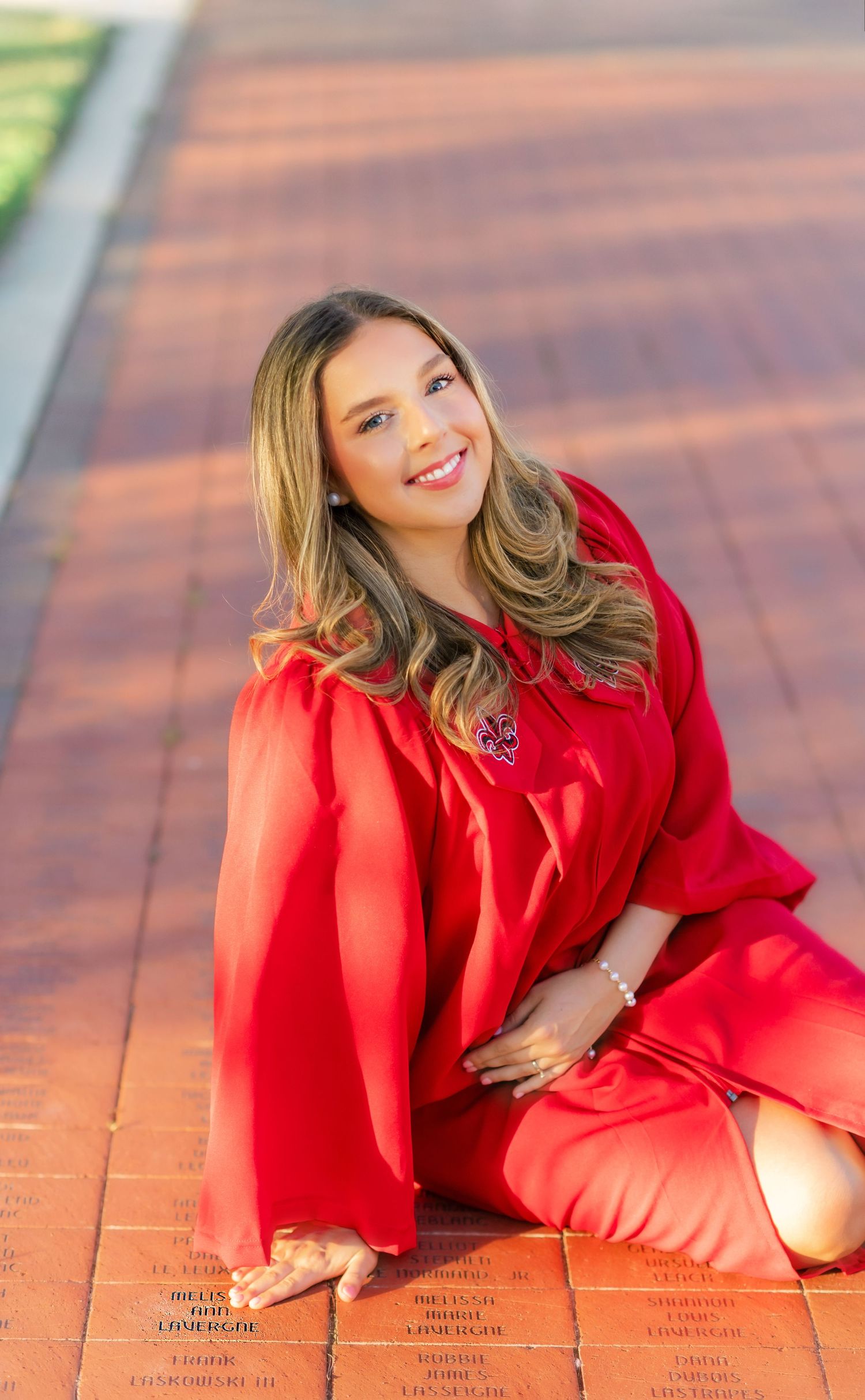 Smiling graduate in red cap and gown sits gracefully on brick walkway with engraved memorial pavers.
