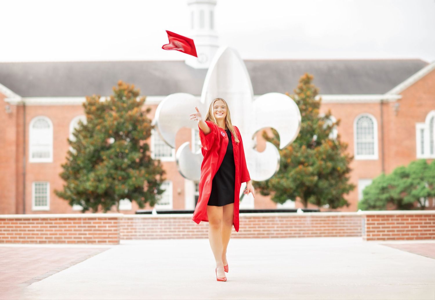 Woman in red graduation gown tossing cap near large floral sculpture outside brick university building.