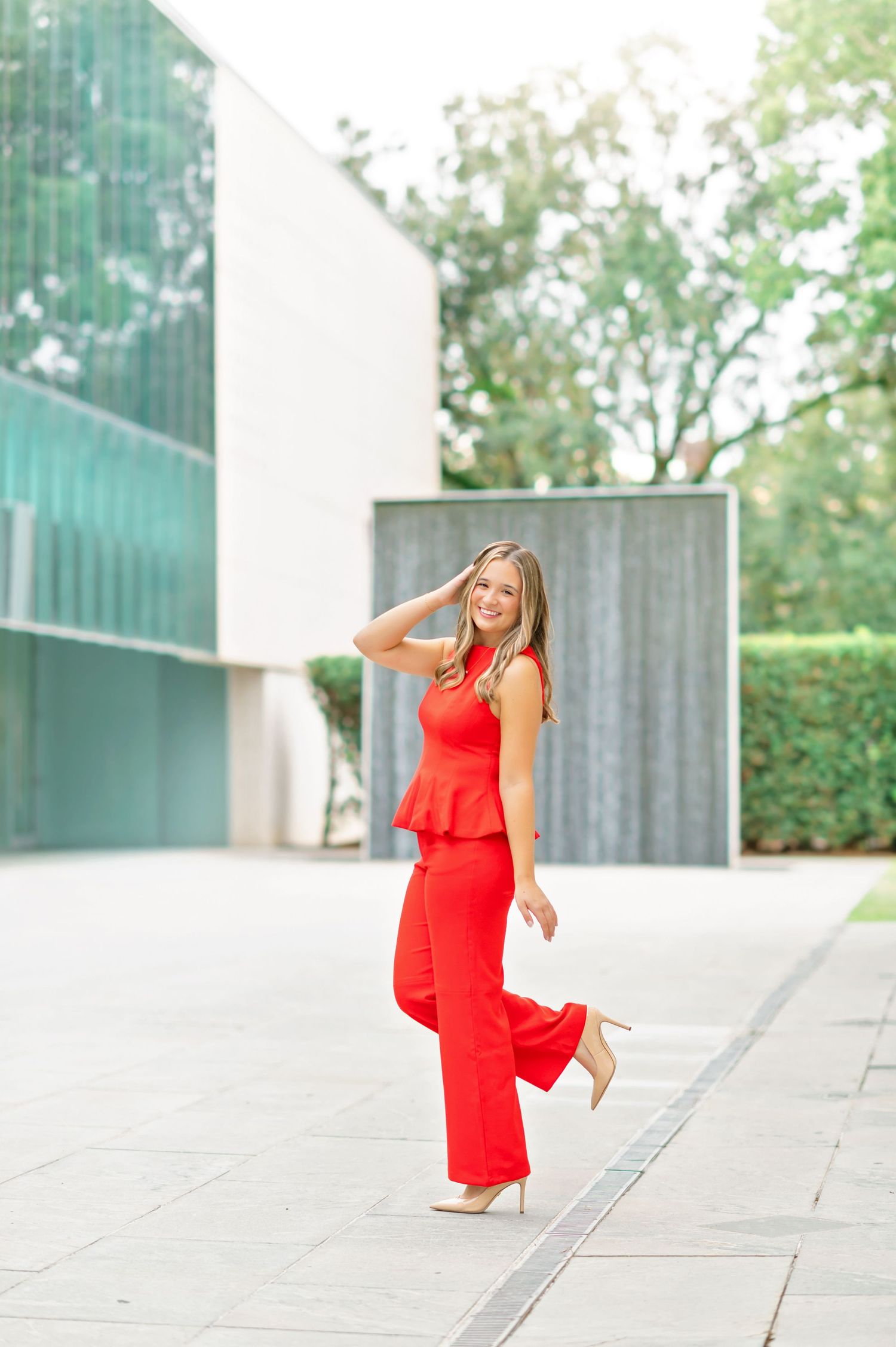Woman in red peplum jumpsuit and nude heels posing outdoors near modern glass building.