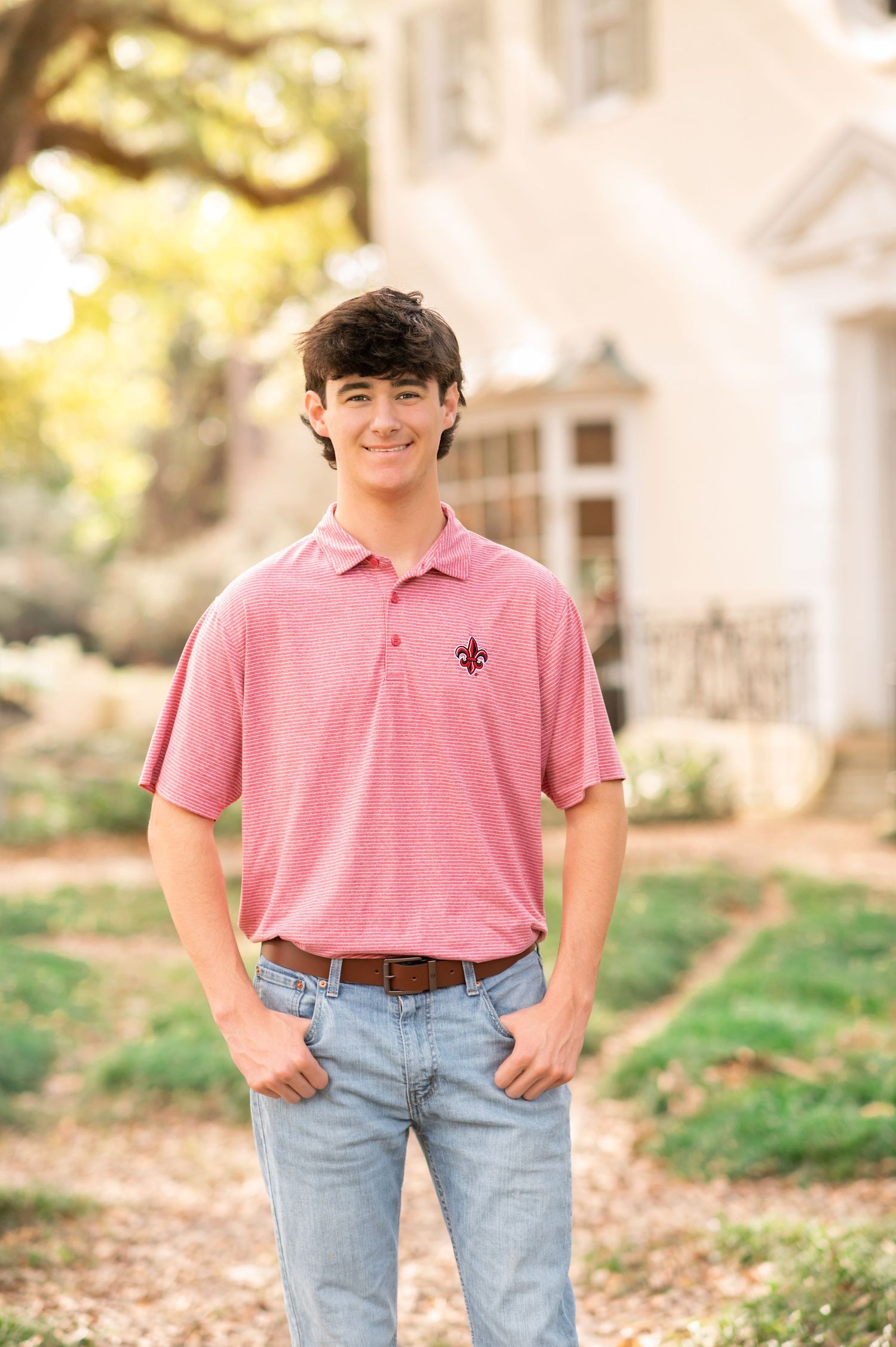 Smiling teenage boy in pink polo shirt and jeans posing outdoors near a white building.