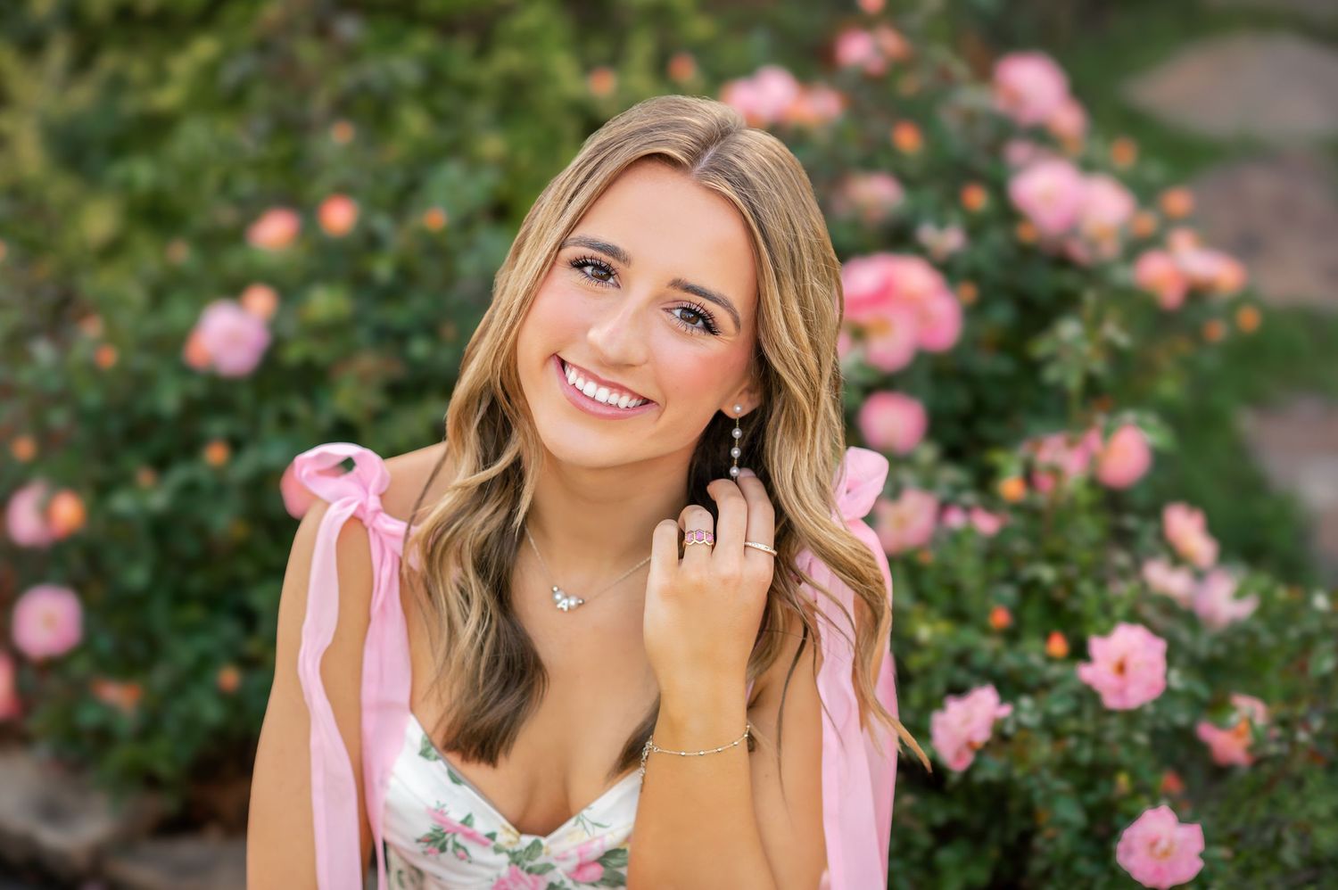 Smiling young woman with blonde hair poses among pink roses wearing a floral top and drop earrings outdoors.