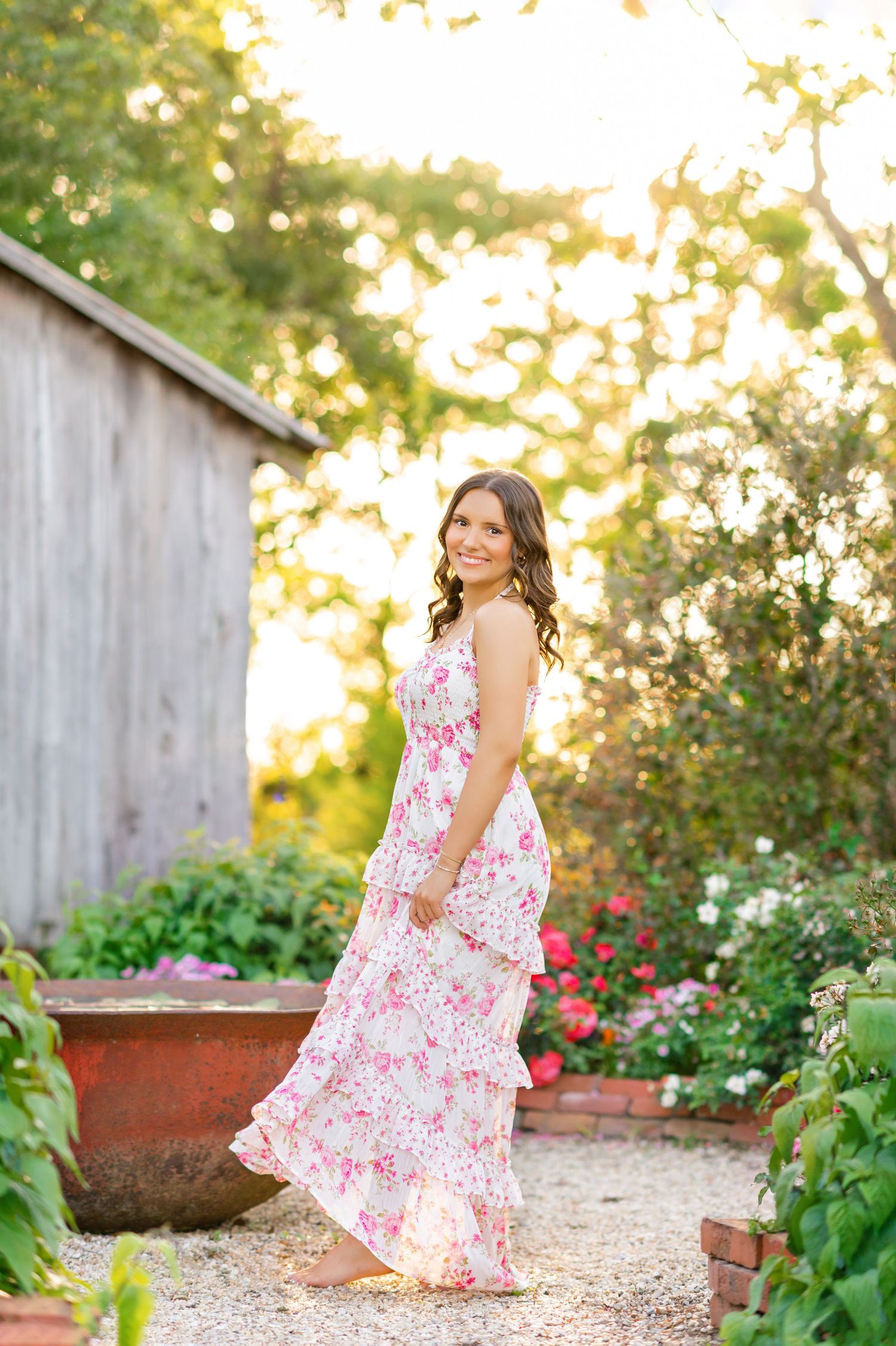Brunette woman in pink floral maxi dress smiling in a garden at golden hour sunset.