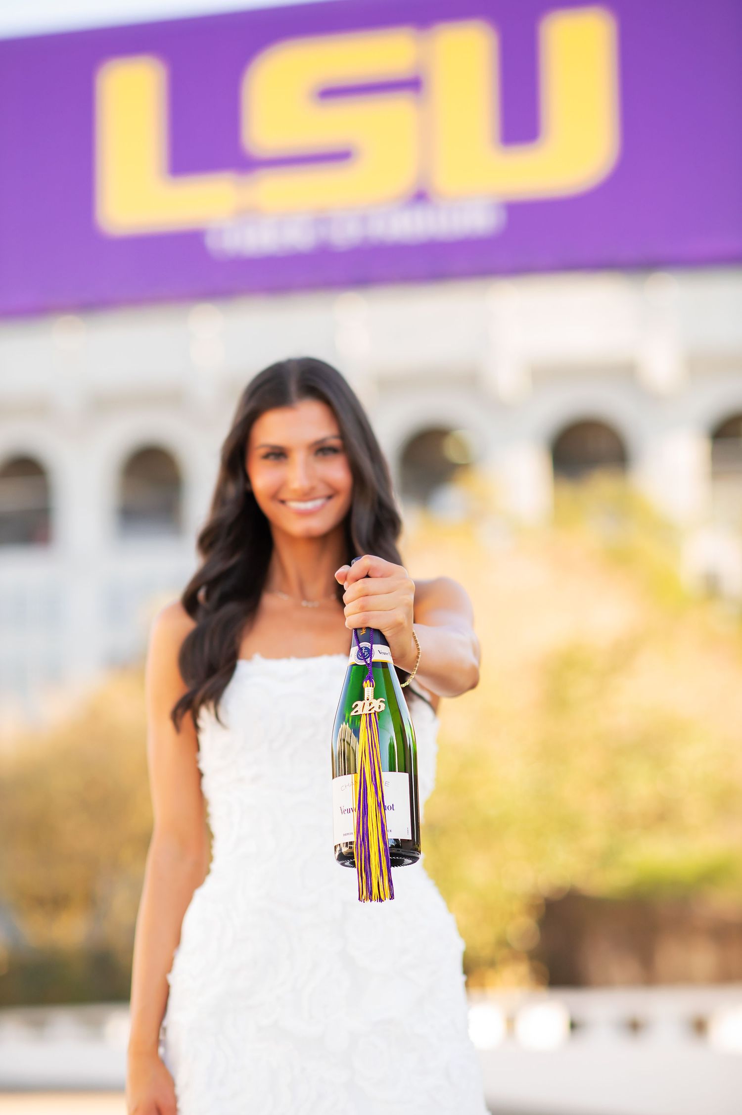 LSU graduate in white dress holds champagne bottle in front of Tiger Stadium.