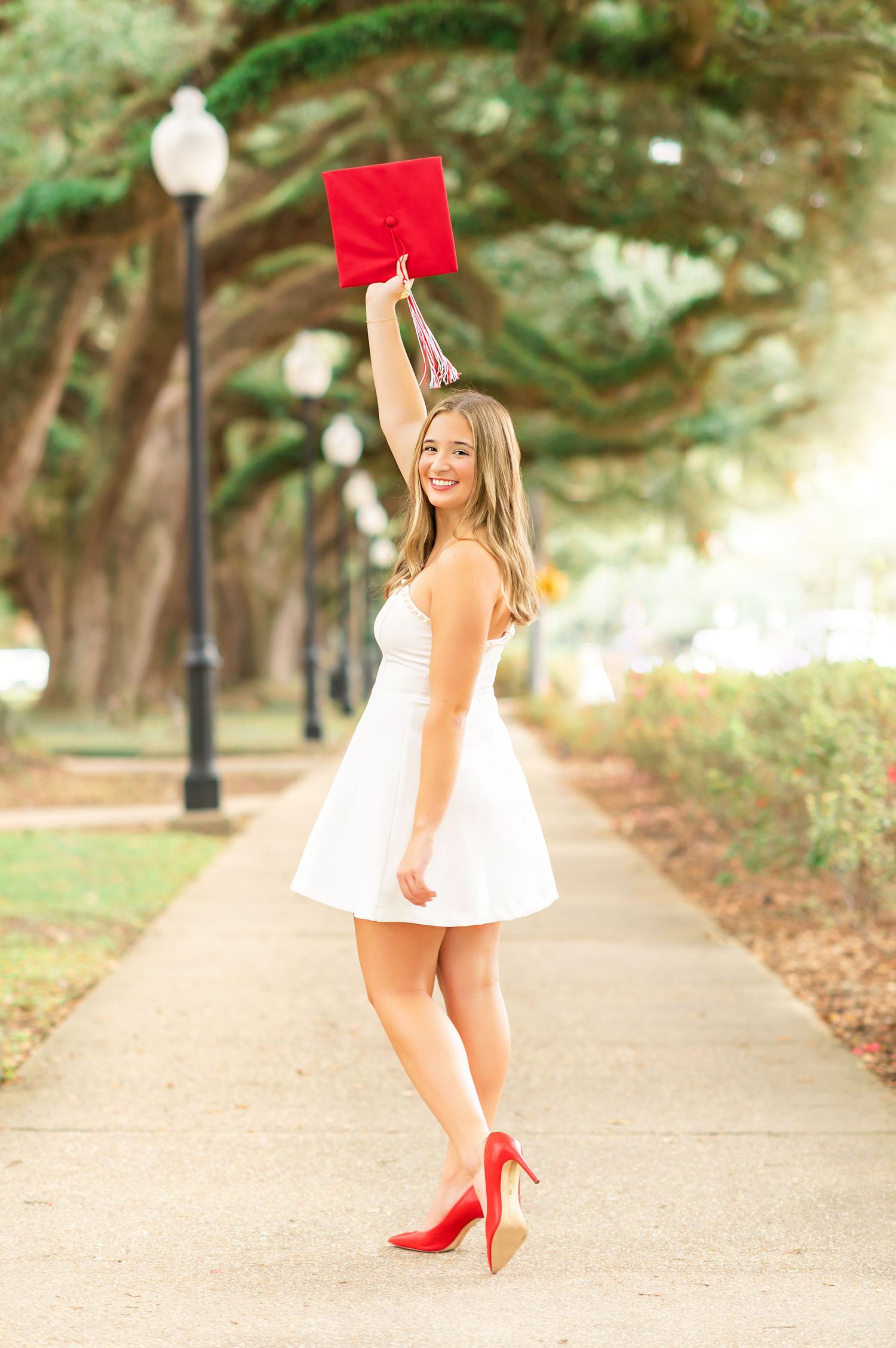 Young woman in white dress and red heels celebrates graduation by tossing red cap on tree-lined walkway.
