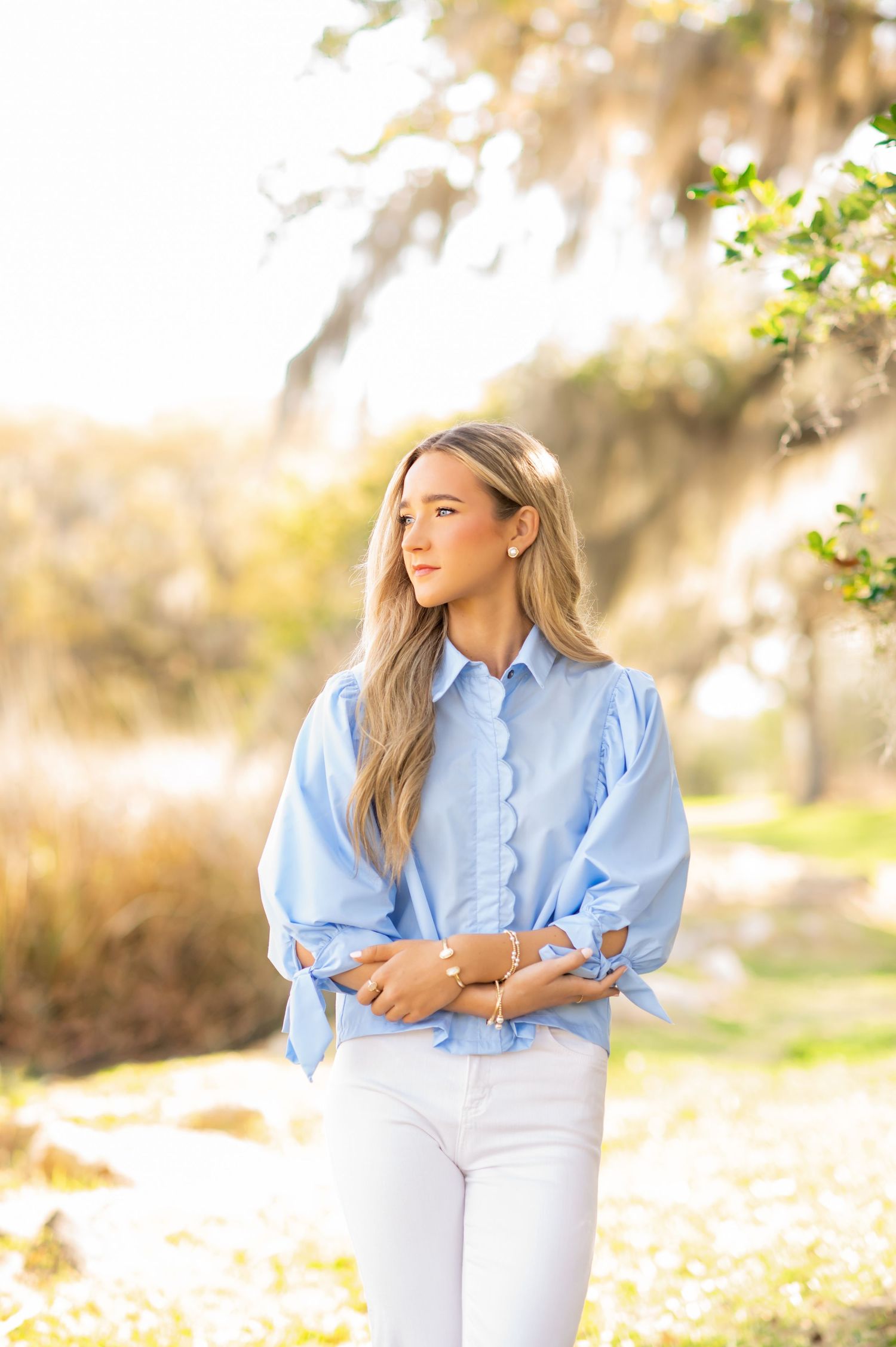 Young woman in light blue scalloped blouse and white jeans posing outdoors among sunlit trees.