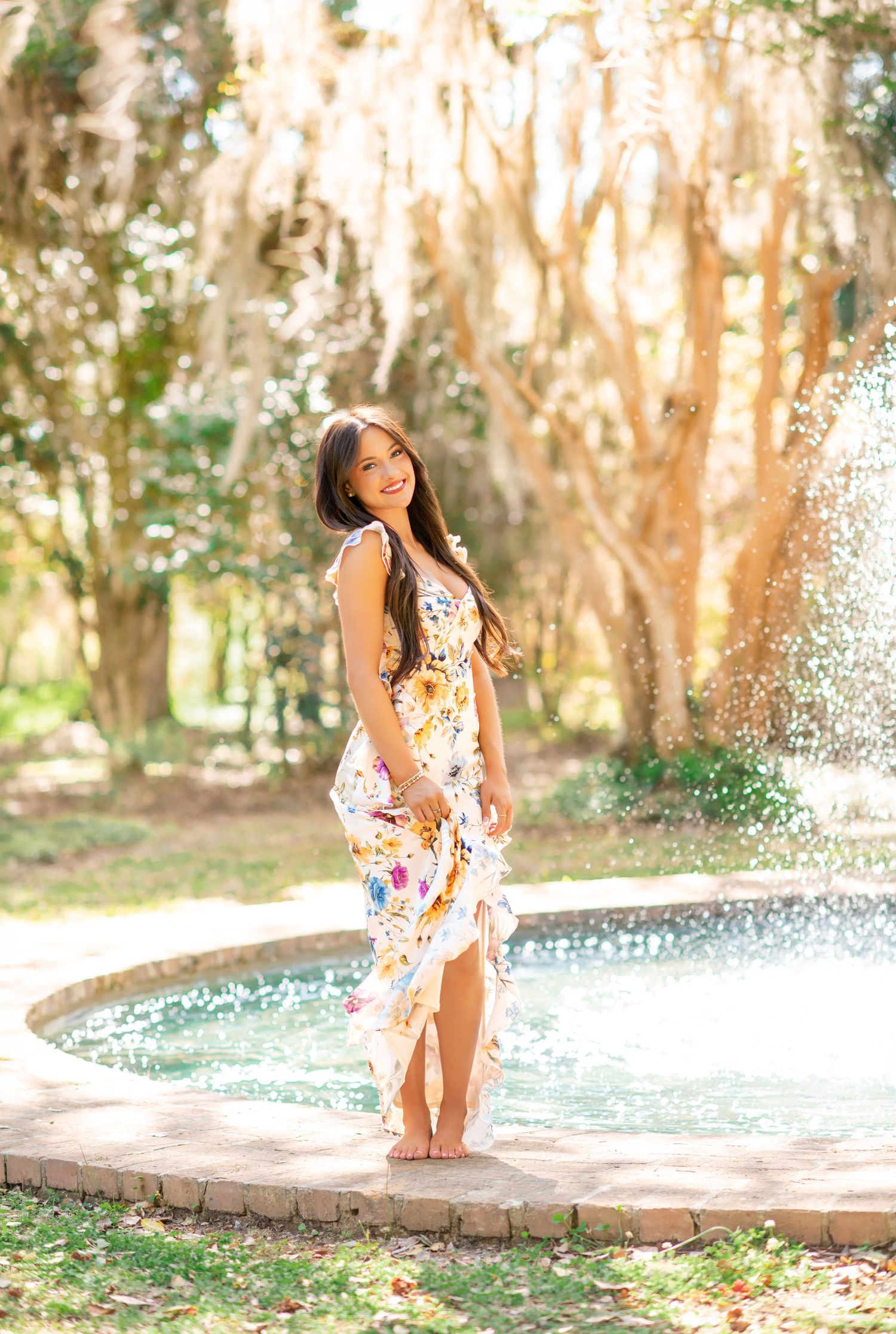 Young woman in floral maxi dress standing barefoot by a garden fountain surrounded by sunlit trees.