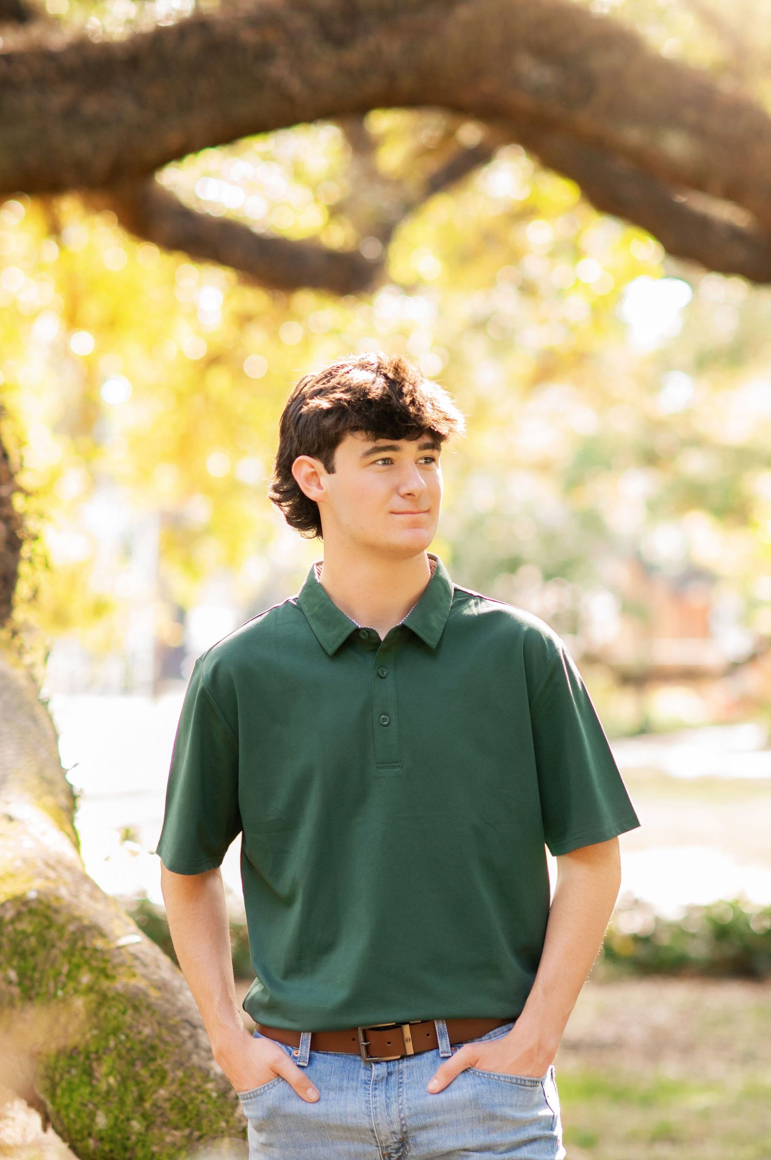 Young man in green polo shirt standing outdoors under autumn trees with hands in pockets.