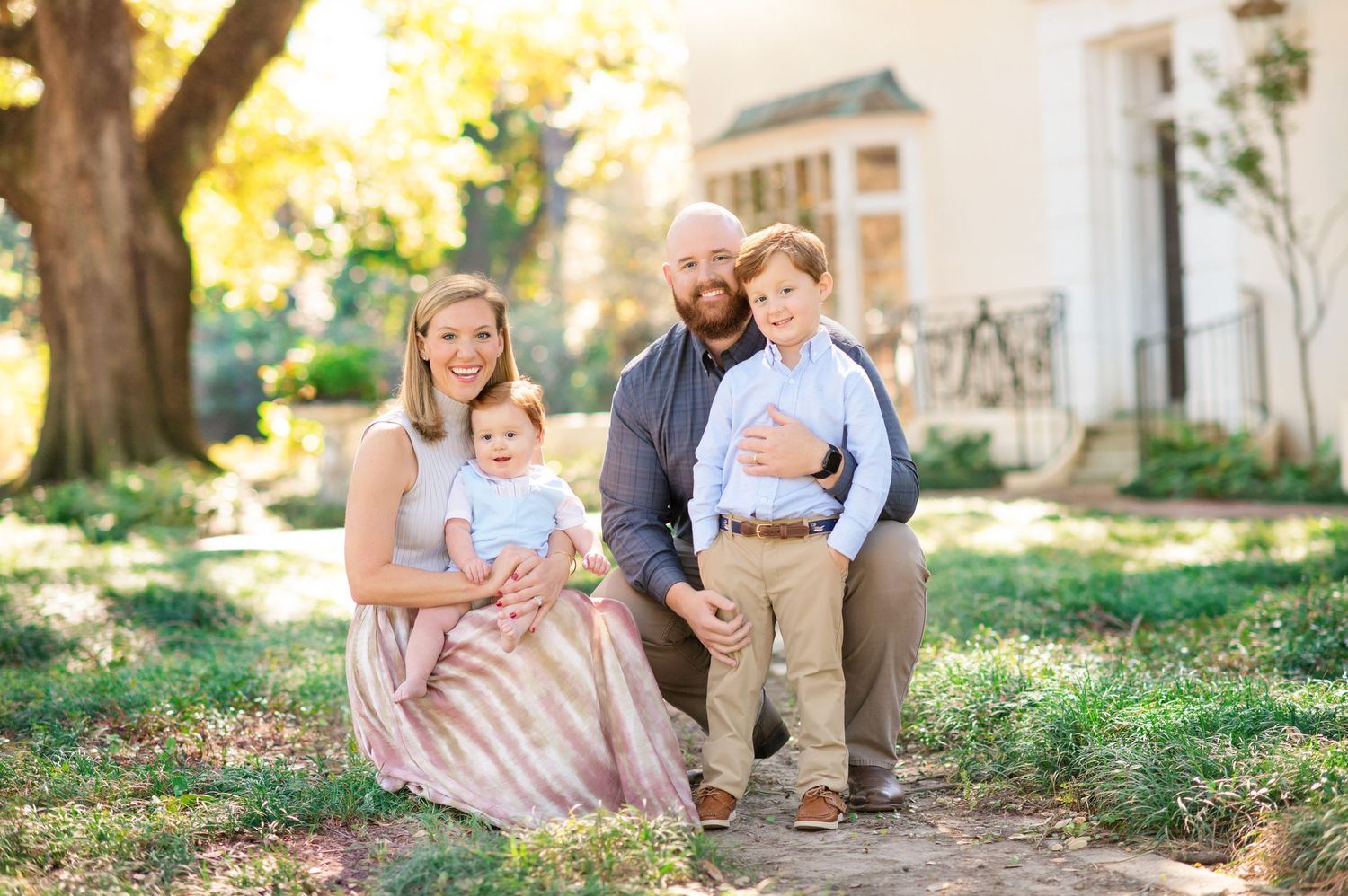 Family of four posing outdoors in golden light, parents holding two young boys near a white building with large trees.