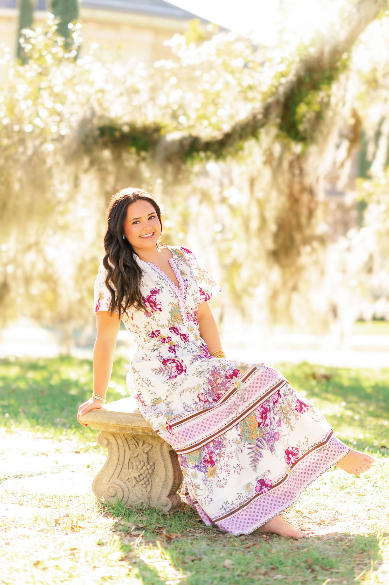 Smiling brunette woman in floral maxi dress sits on stone bench under Spanish moss in golden sunlight.